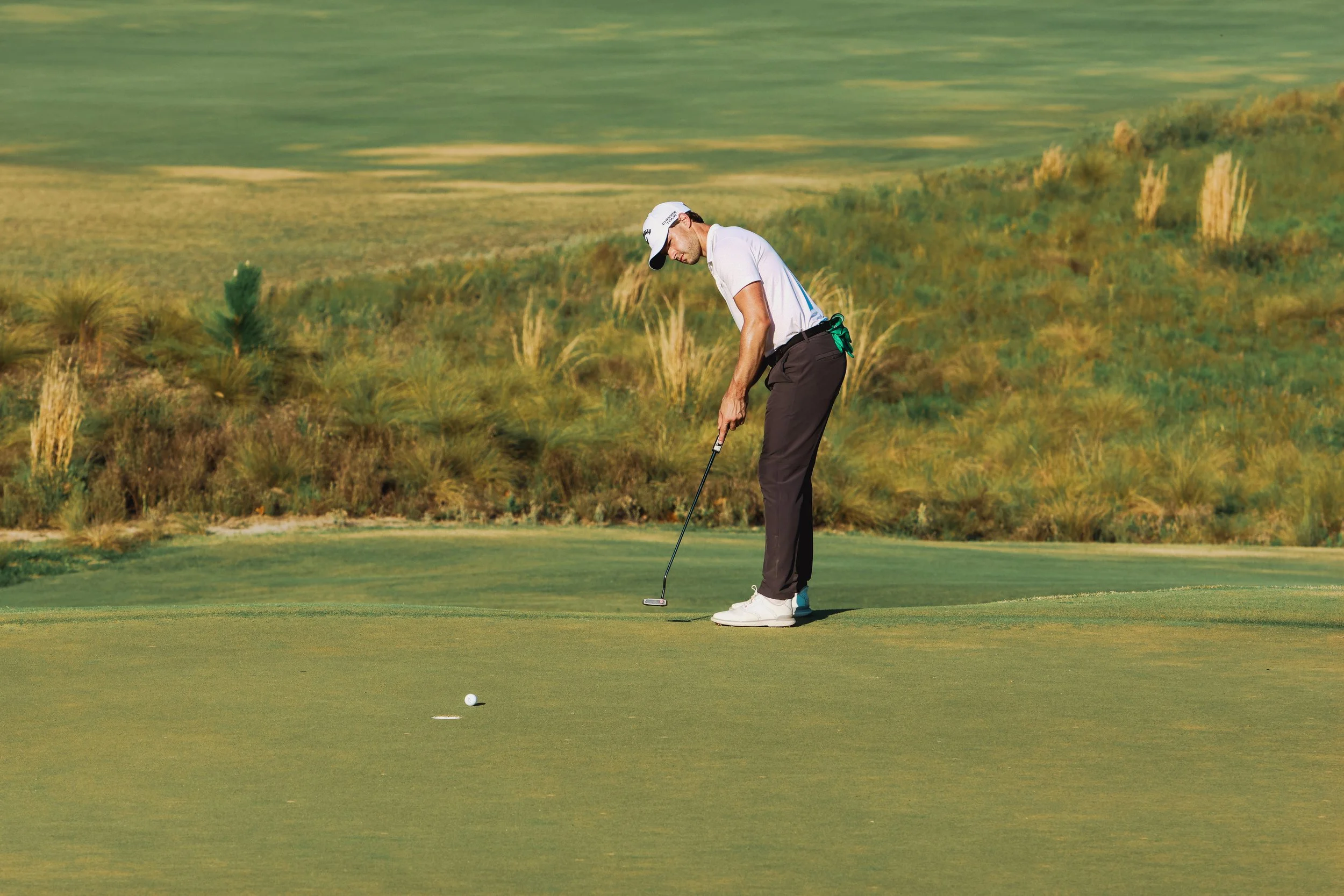 A man wearing a white golf cap, white polo shirt, and black pants prepares to putt a golf ball on a green golf course during daytime.