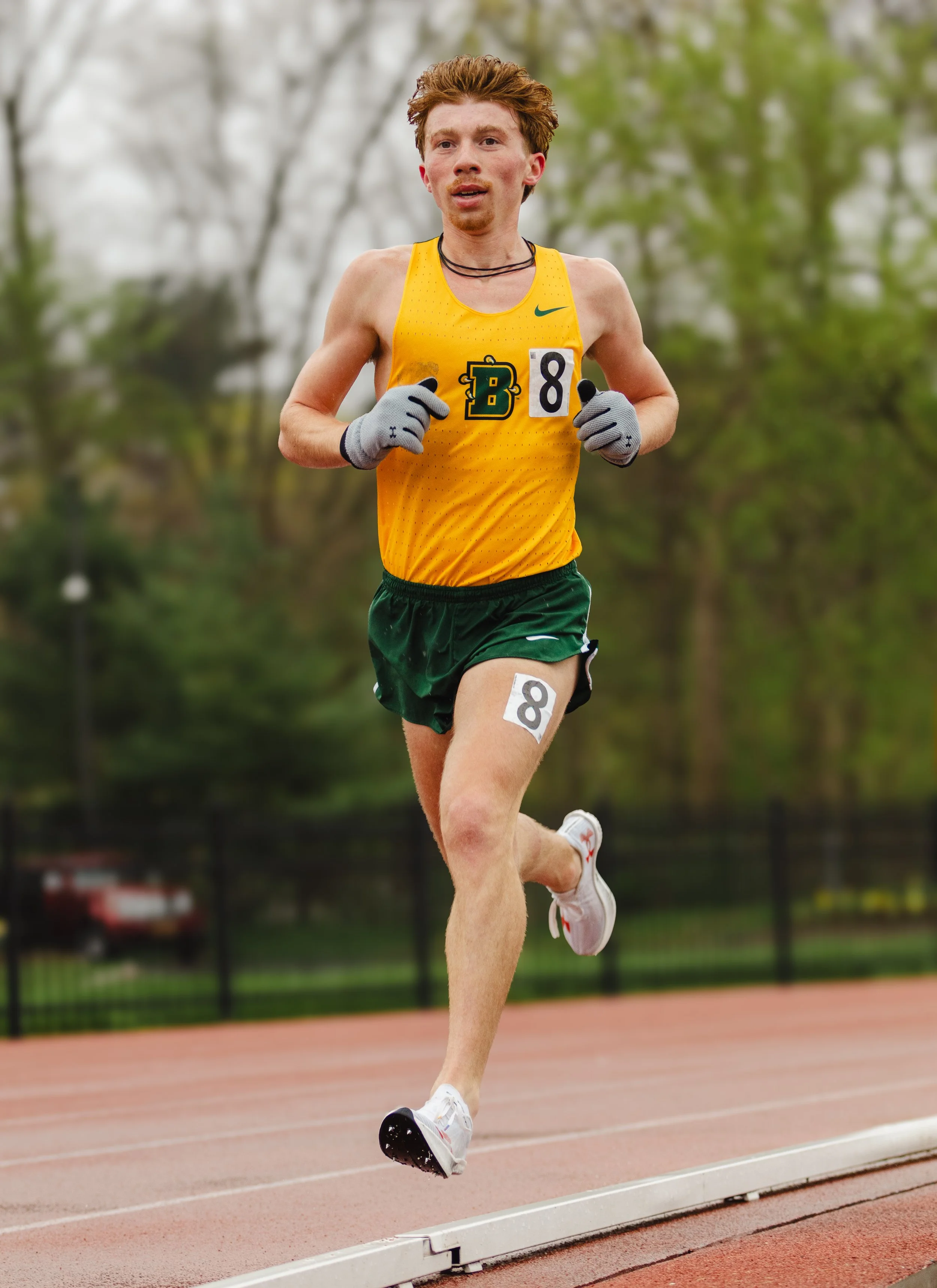 A male athlete in a yellow tank top and dark green shorts running on a track with a focused expression.