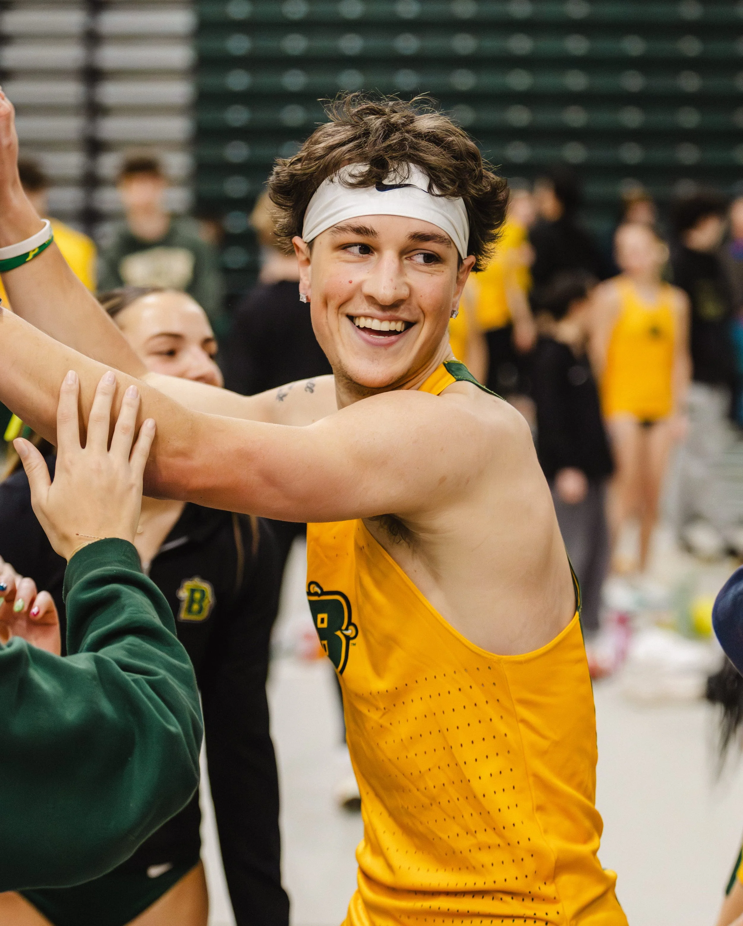 A smiling male athlete with brown hair, wearing a white headband and a yellow sports jersey from SUNY Brockport, celebrating with teammates in a indoor track gymnasium.