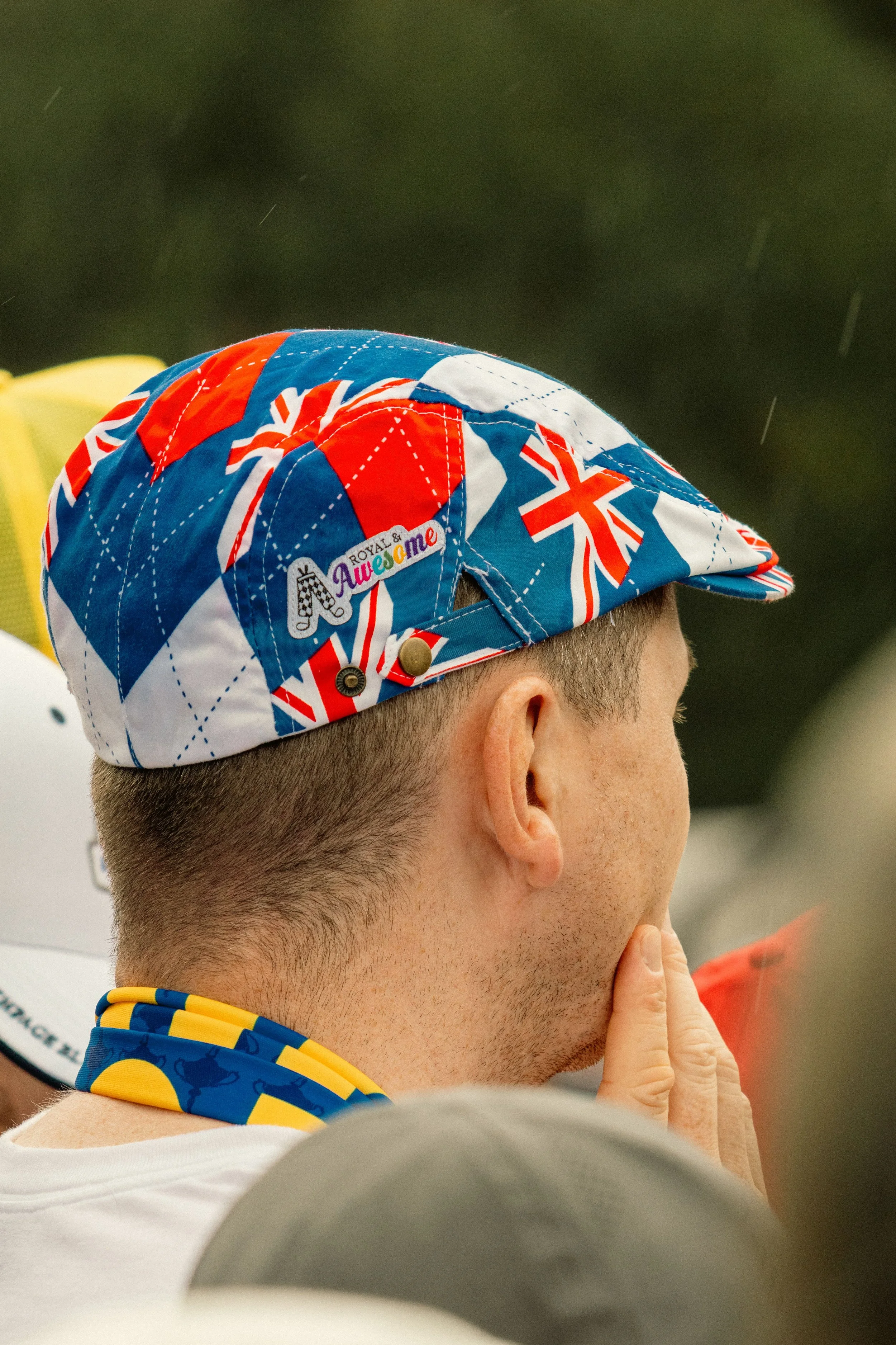 Person wearing a colorful cap with British flags and a sticker that says "Royal & Awesome", with a yellow and blue neck gaiter, in a crowd during an outdoor event.
