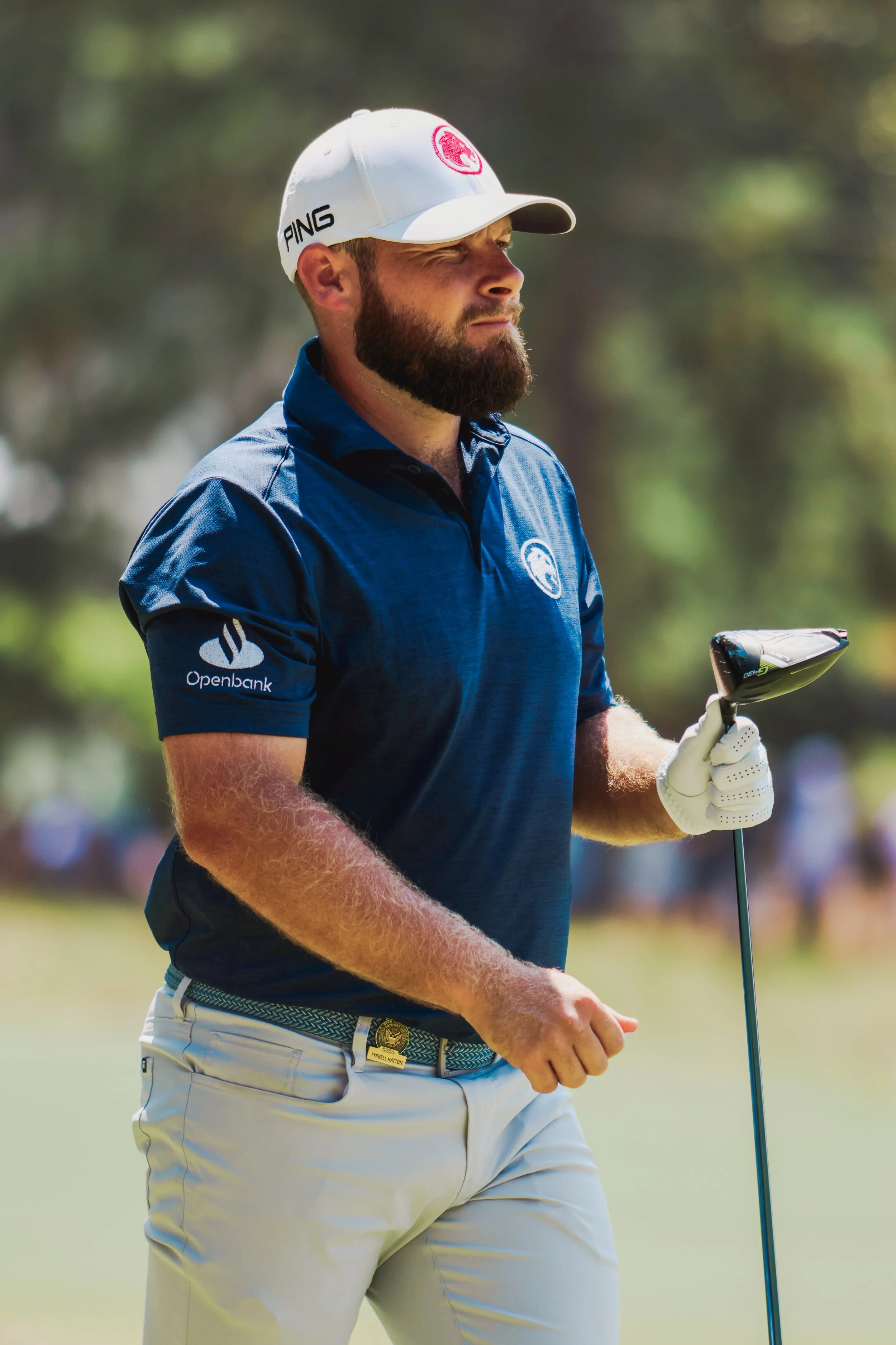 A man with a beard, wearing a white cap and blue shirt, is holding a golf club on a golf course.