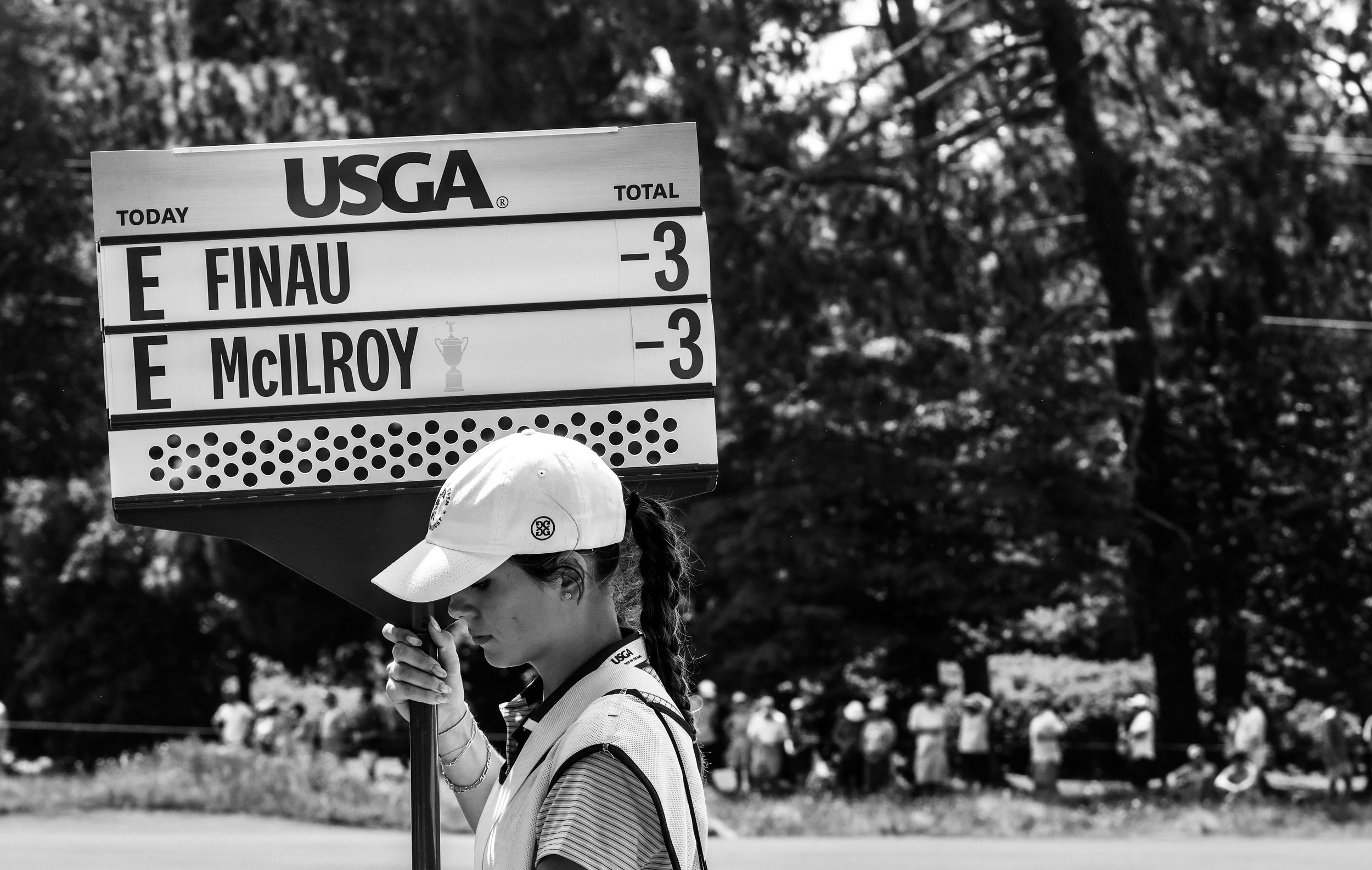 A female golf caddie holding a leaderboard at a golf tournament, with trees and spectators in the background.