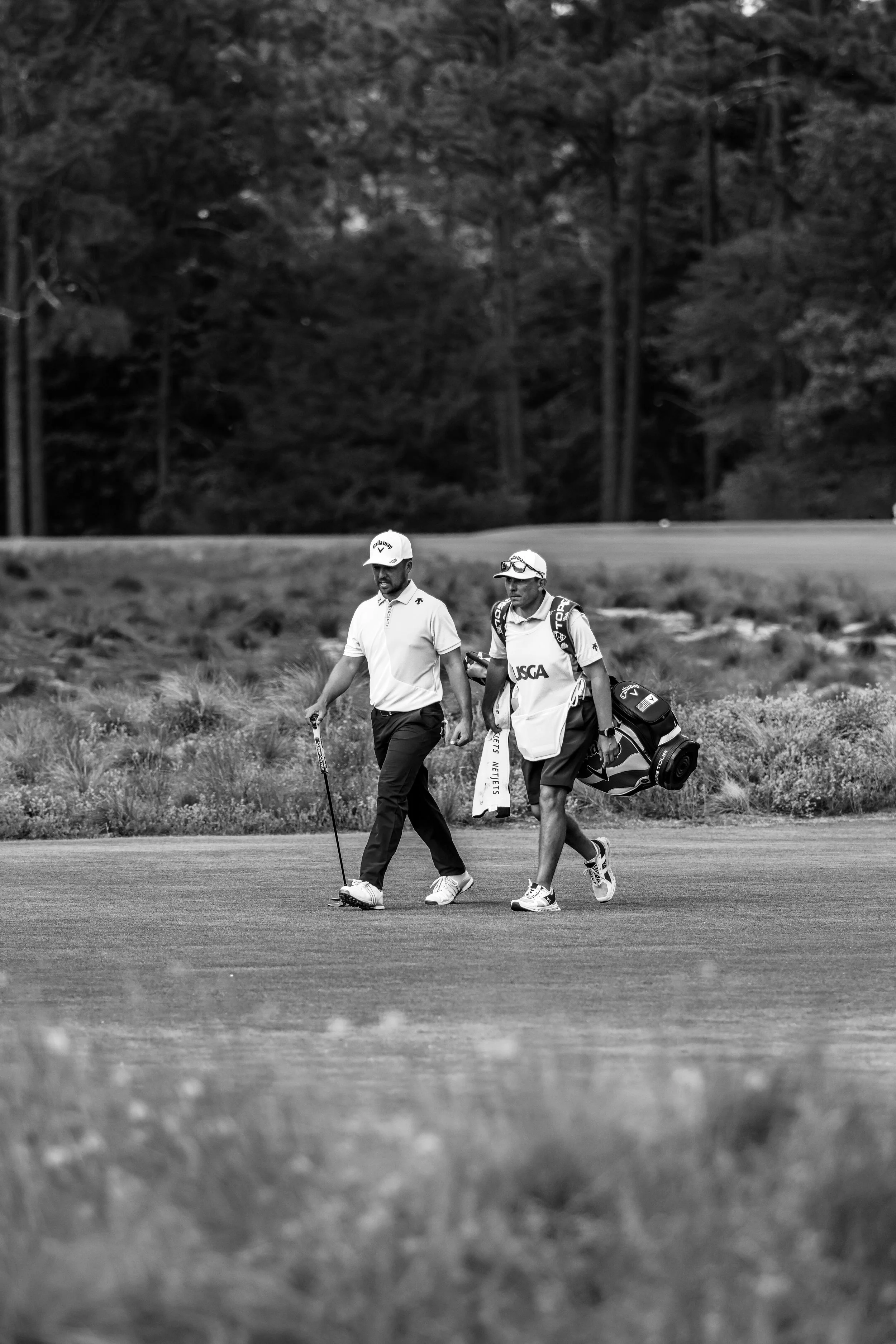 Two men walking on a golf course with golf bags, trees in the background, in black and white.