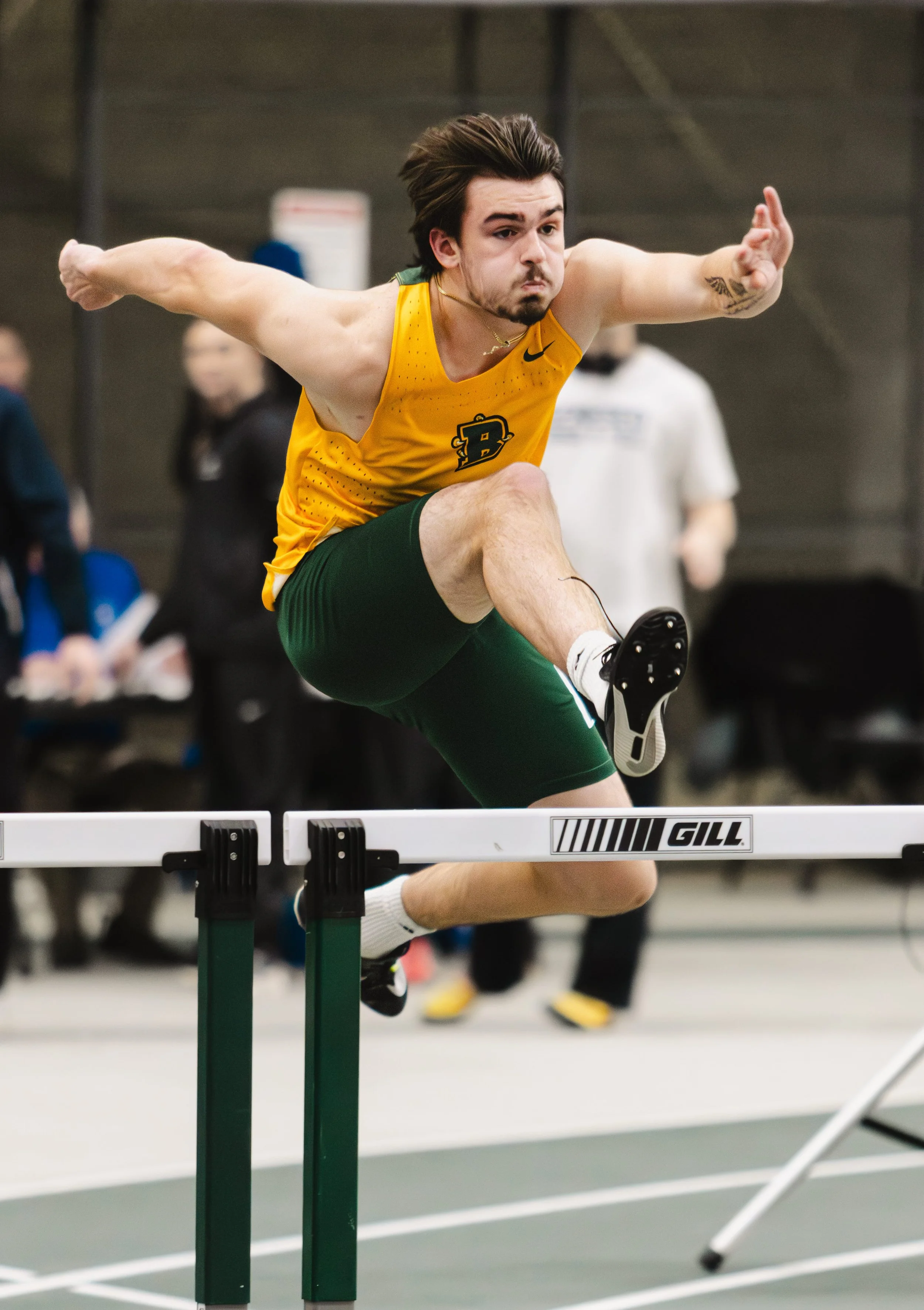 Male athlete from SUNY Brockport in yellow jersey and green shorts clearing a hurdle in an indoor track event.