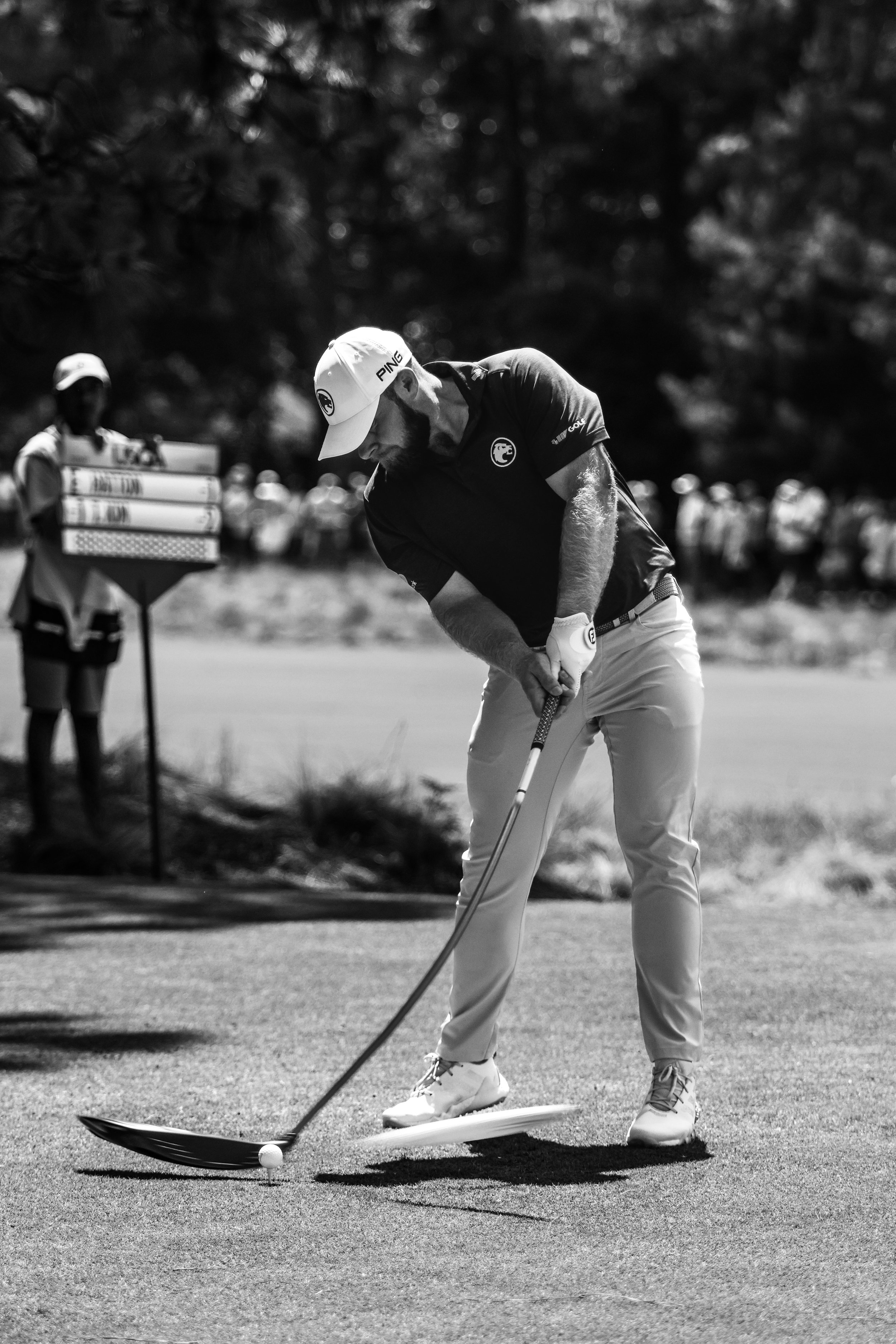 Black and white photo of a golfer about to putt on a golf course. The golfer is in focus, wearing a cap and polo shirt, with a group of spectators in the background along with a caddy holding a golf bag.