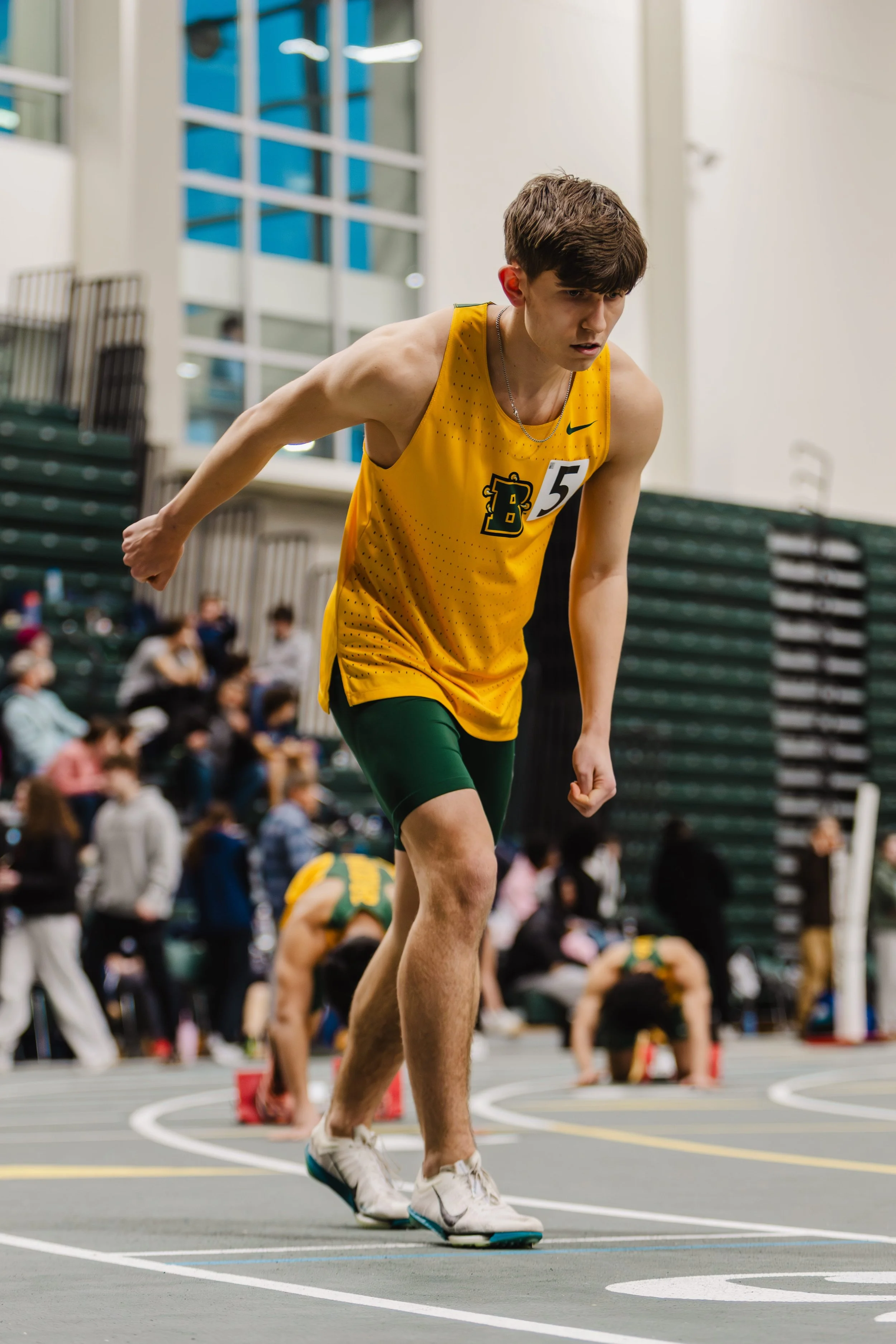 Young male track athlete in yellow jersey and black shorts from SUNY Brockport starting a race on indoor track.