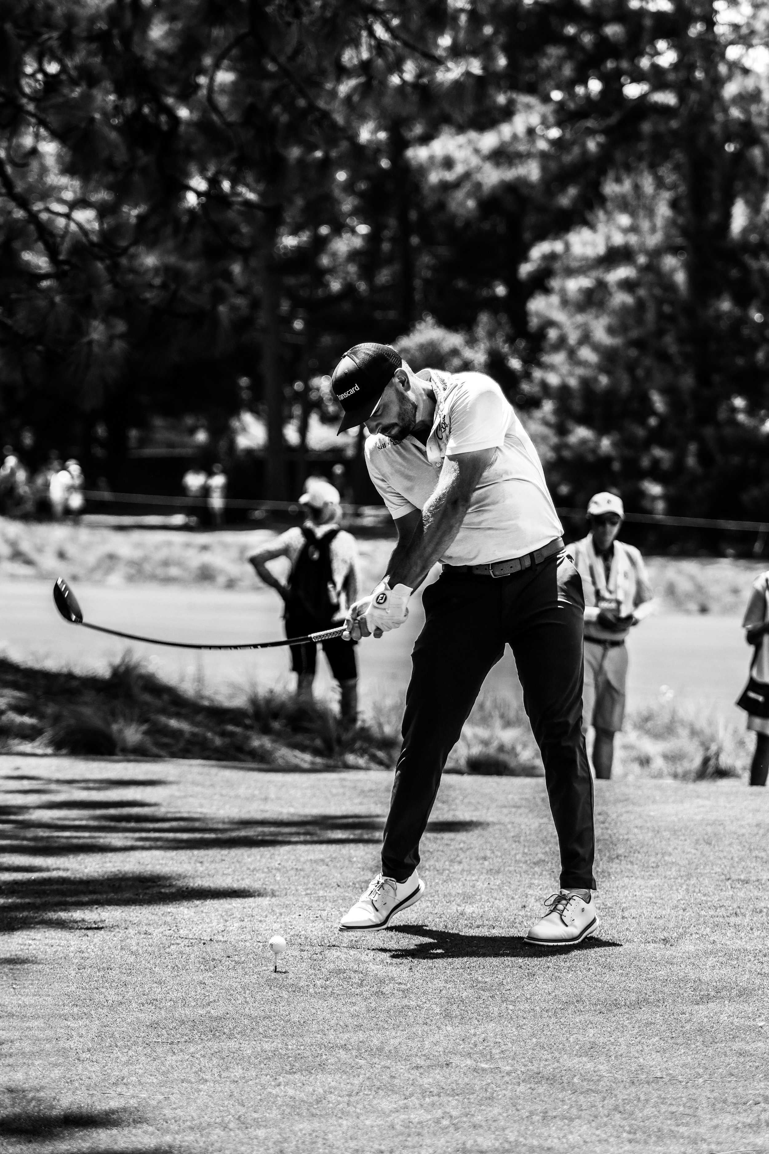 A man playing golf on a golf course, preparing to hit a golf ball with a club, with several people observing in the background on a sunny day.