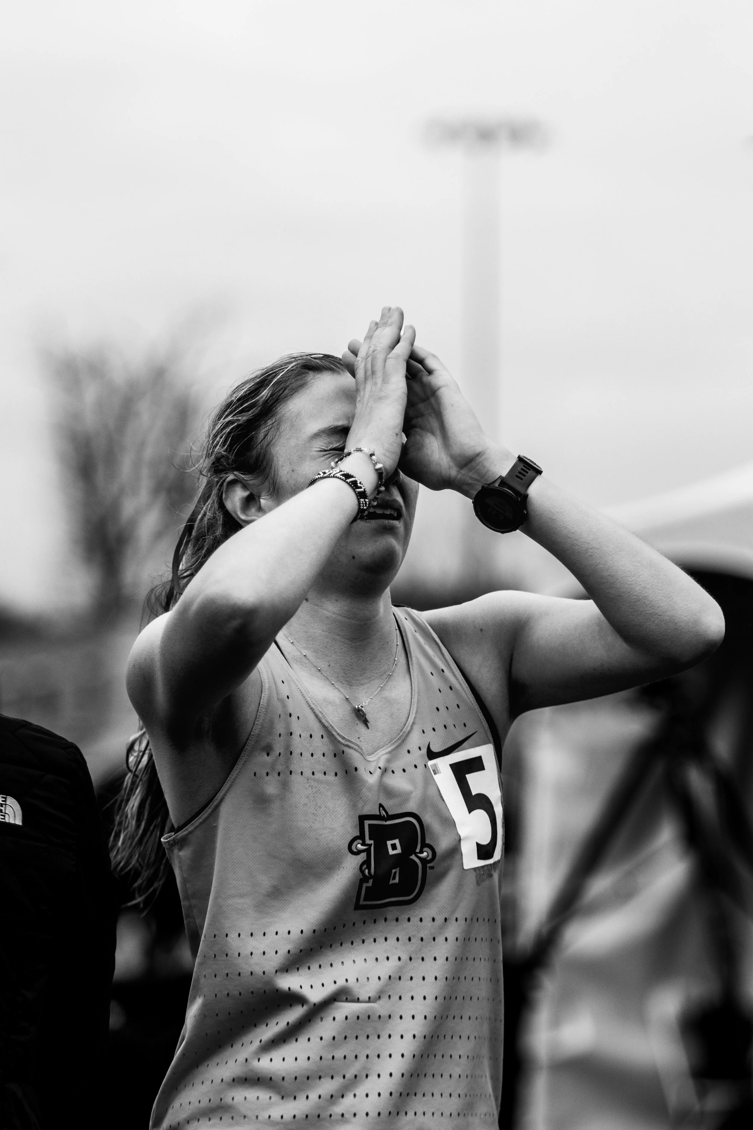 A young woman smiling and celebrating after a running race, wearing athletic gear with the number 5 pinned to her shirt, with her hands pressed together in a gesture of gratitude or prayer, outdoors on a cloudy day.