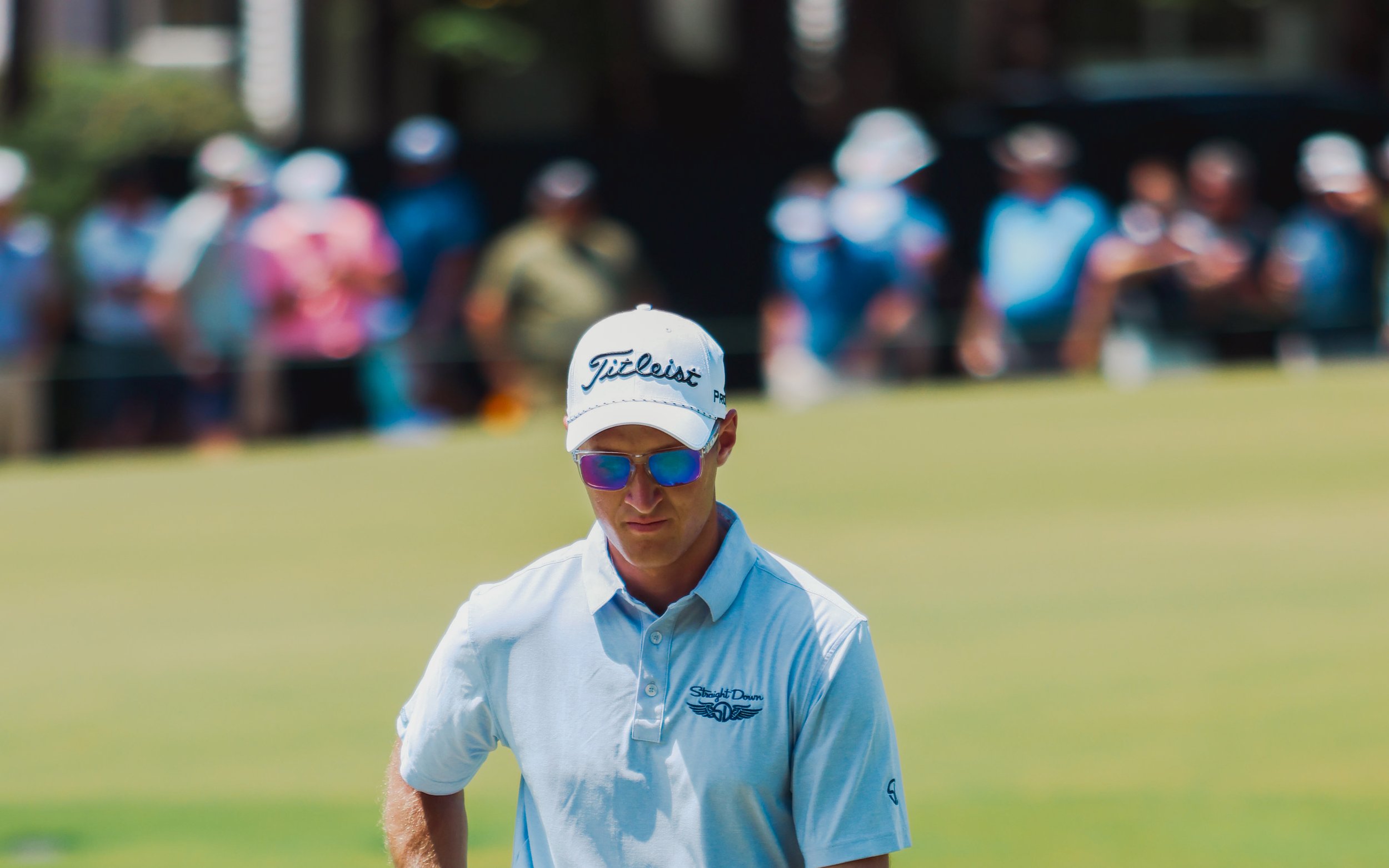 A man in a white golf shirt and white baseball cap with the Titleist logo, wearing sunglasses, standing on a golf course with a crowd of spectators blurred in the background.