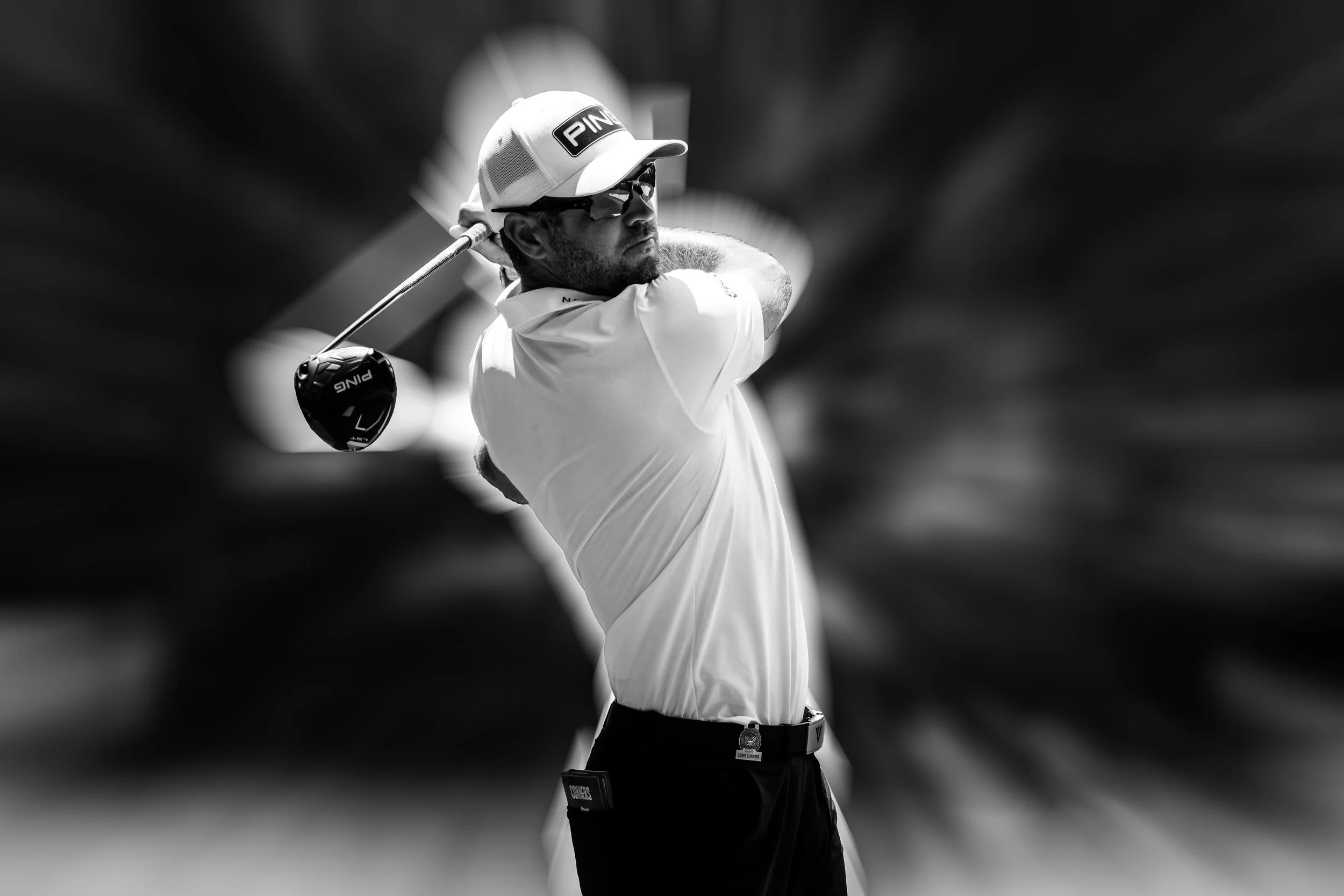 Black and white photo of a male golfer wearing a cap and sunglasses, swinging a golf club.