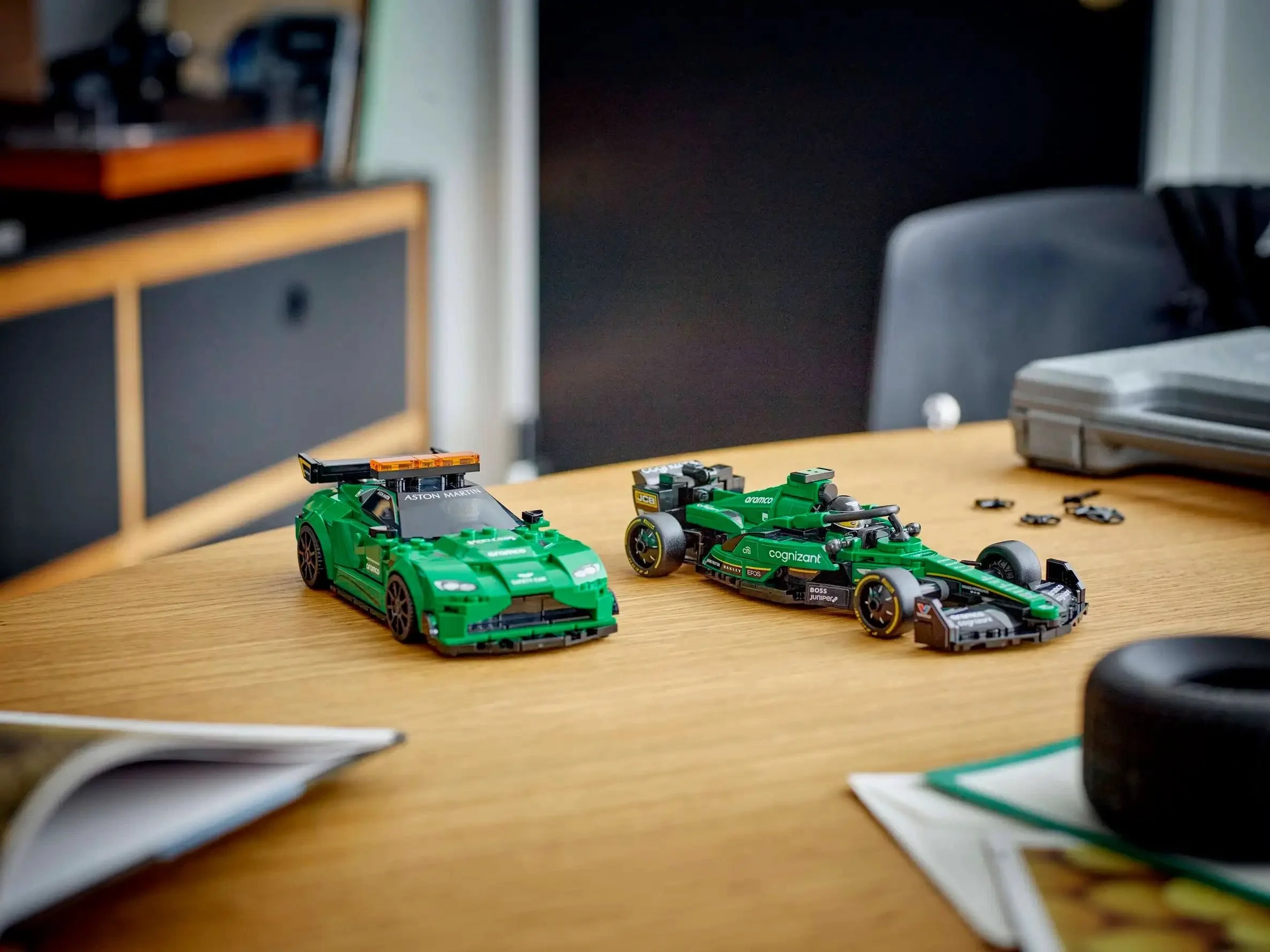 Two green LEGO race cars on a wooden table, one resembling a Formula 1 car and the other a sports car, with some loose LEGO pieces nearby.