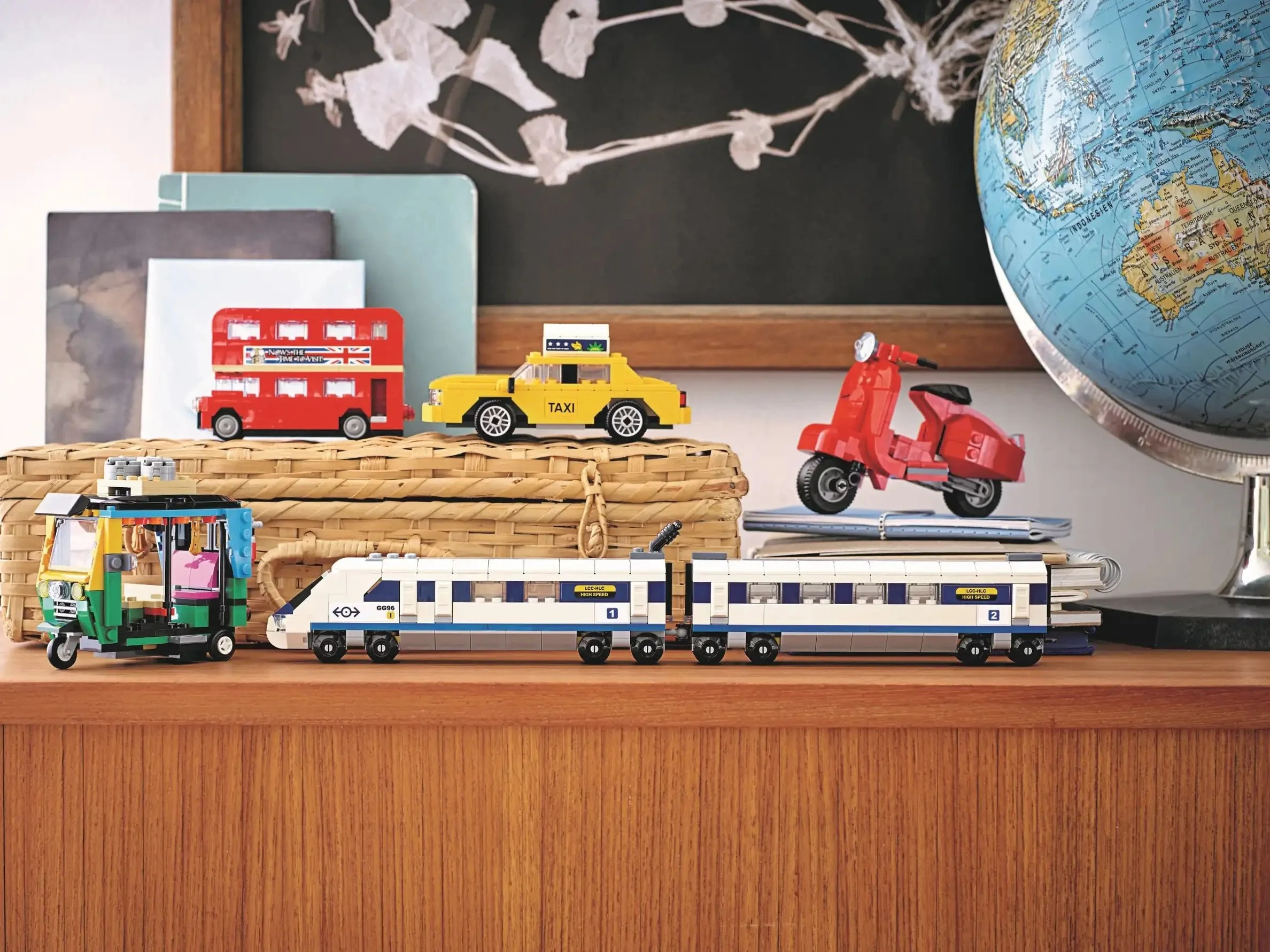 Model vehicles including a red double-decker bus, yellow taxi, red scooter, green shop vehicle, and a white and blue train set, arranged on a wooden surface with books and a globe in the background.