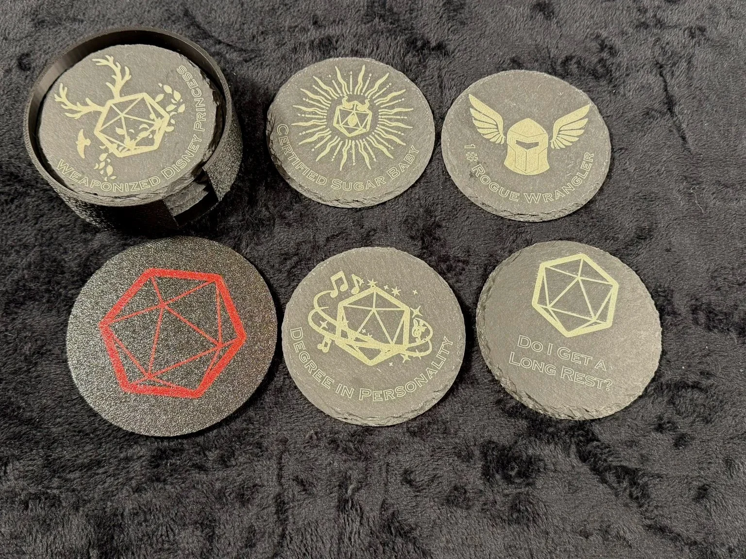 Six round coasters with various Dungeons and Dragons themed designs and phrases, arranged on a black slate surface, with some coasters stacked in a holder.