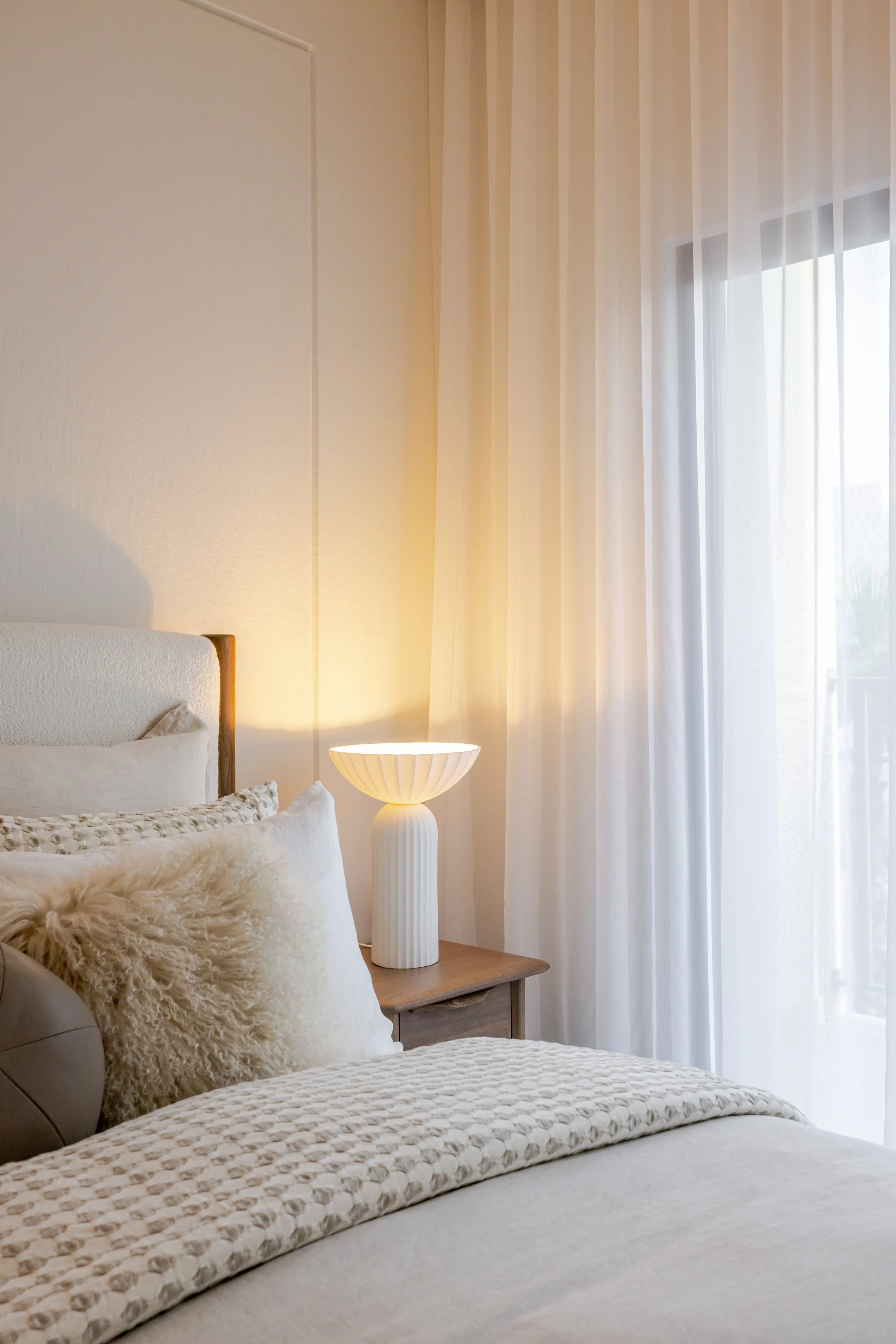 A cozy bedroom with a bed featuring textured pillows, a bedside table with a modern white table lamp, and sheer white curtains allowing natural light to fill the room.