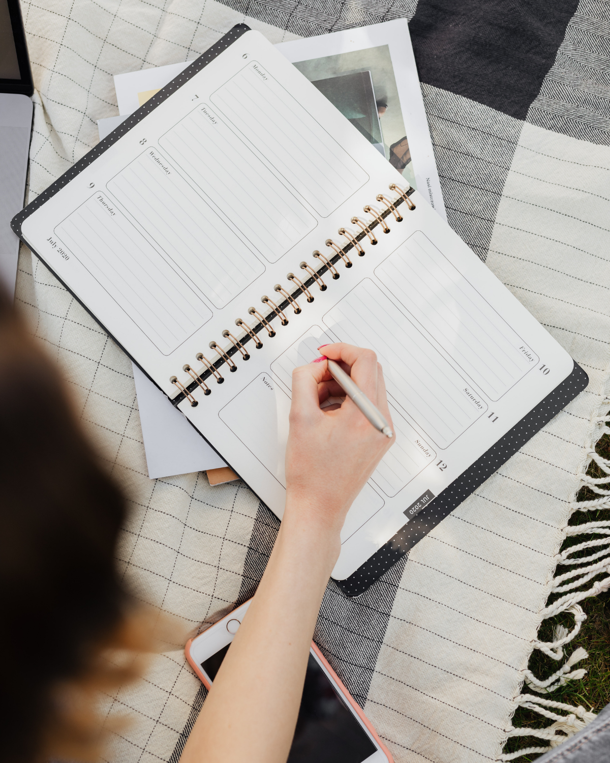 A person writing in a weekly planner notebook on a checkered cloth, with a smartphone and a laptop nearby.