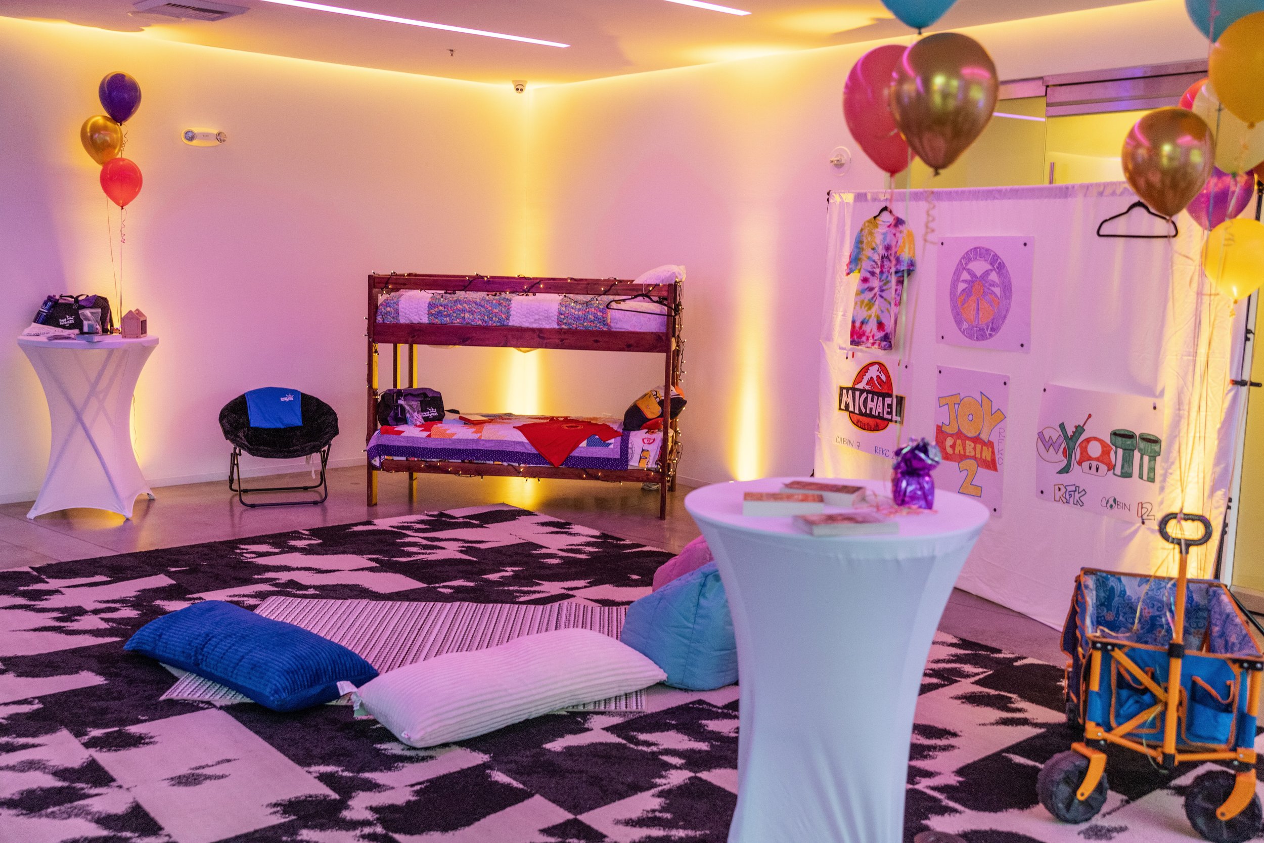 Party room decorated with colorful balloons, a bunk bed with a quilt, pillows on the floor, and a display with toys and artwork, illuminated by warm lighting.