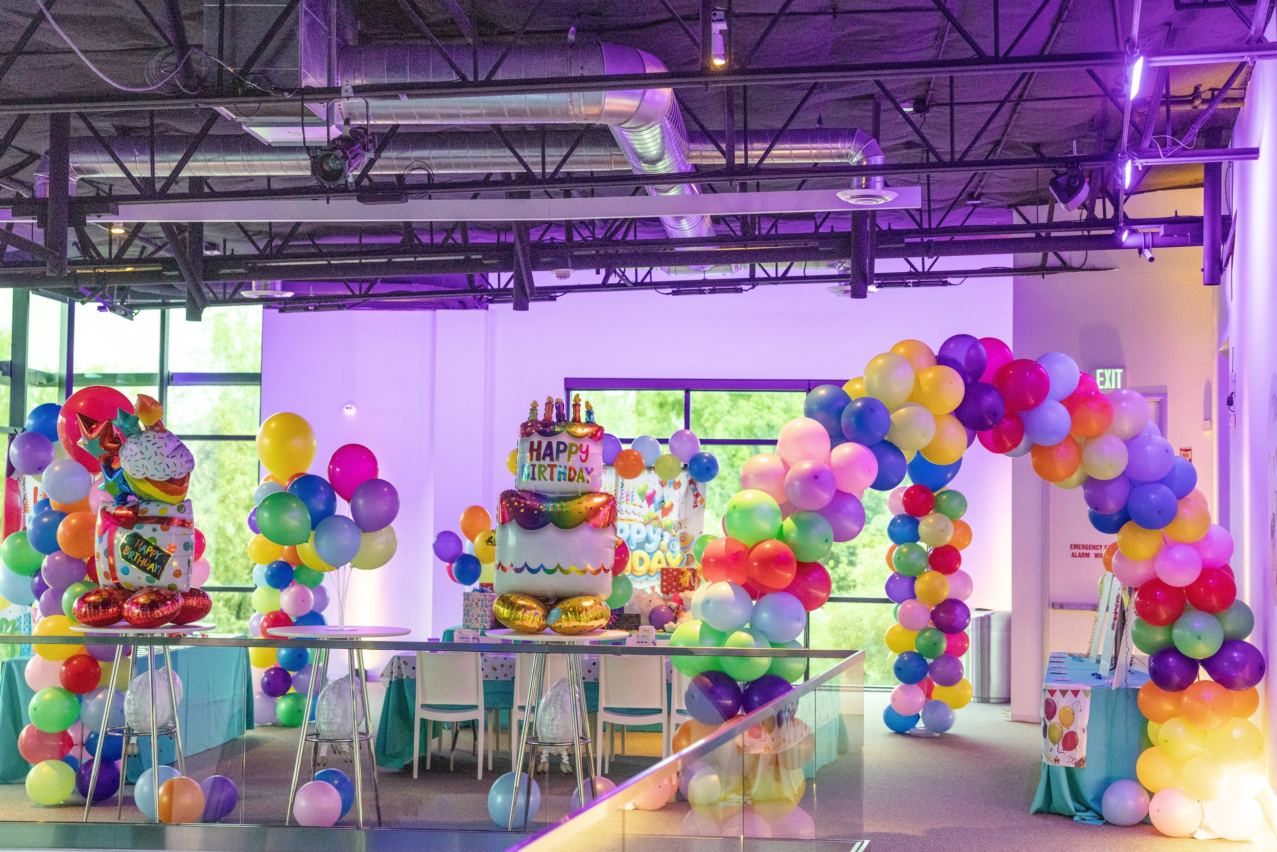 Decorations for a birthday party featuring numerous multicolored balloons, a large birthday cake with candles, and birthday-themed balloons and banners, all set inside a modern room with large windows and purple lighting.