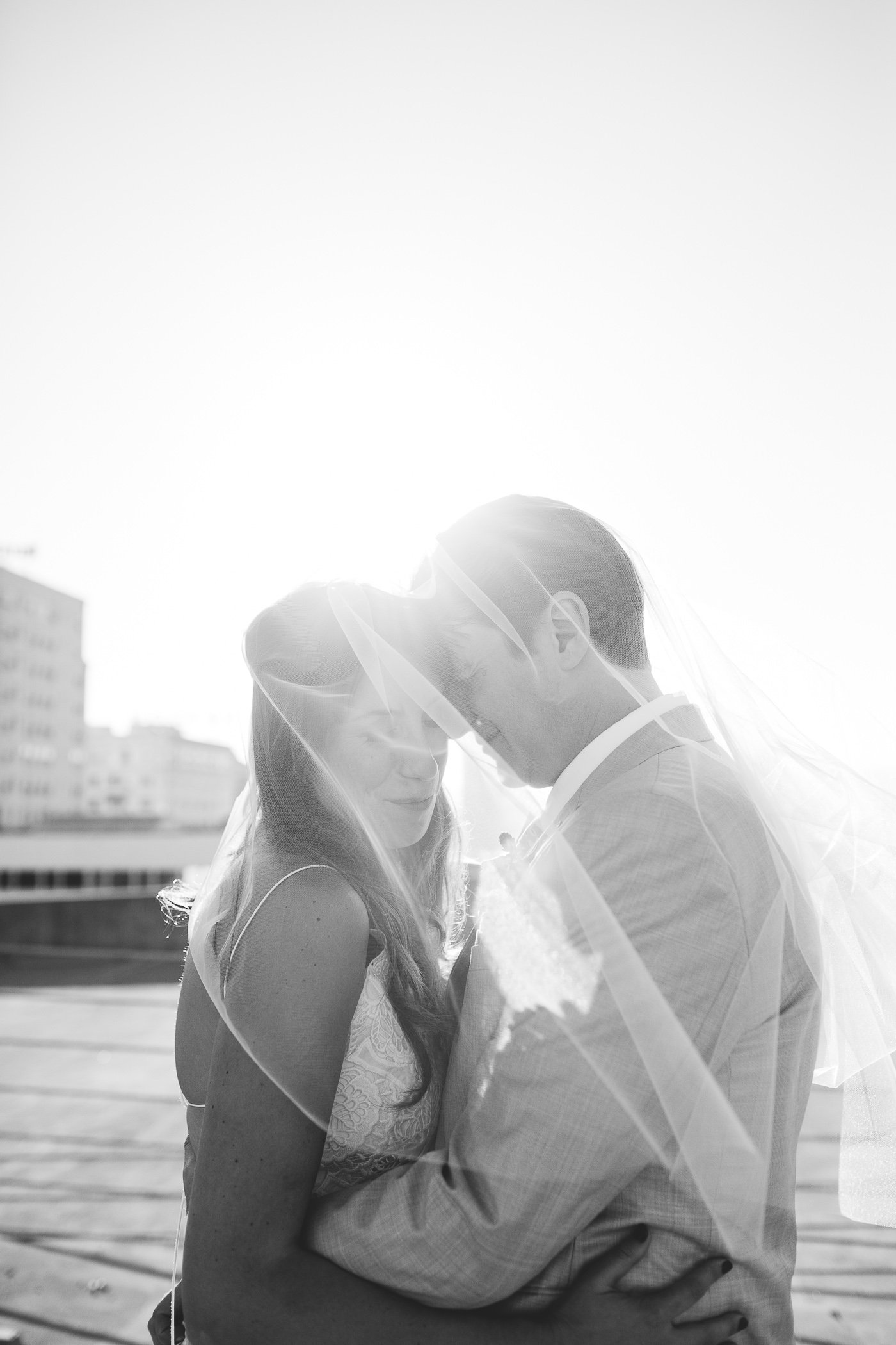 A black and white photo of a couple embracing and kissing on a rooftop with city buildings in the background, with the bride wearing a lace wedding dress and the groom in a light-colored suit.