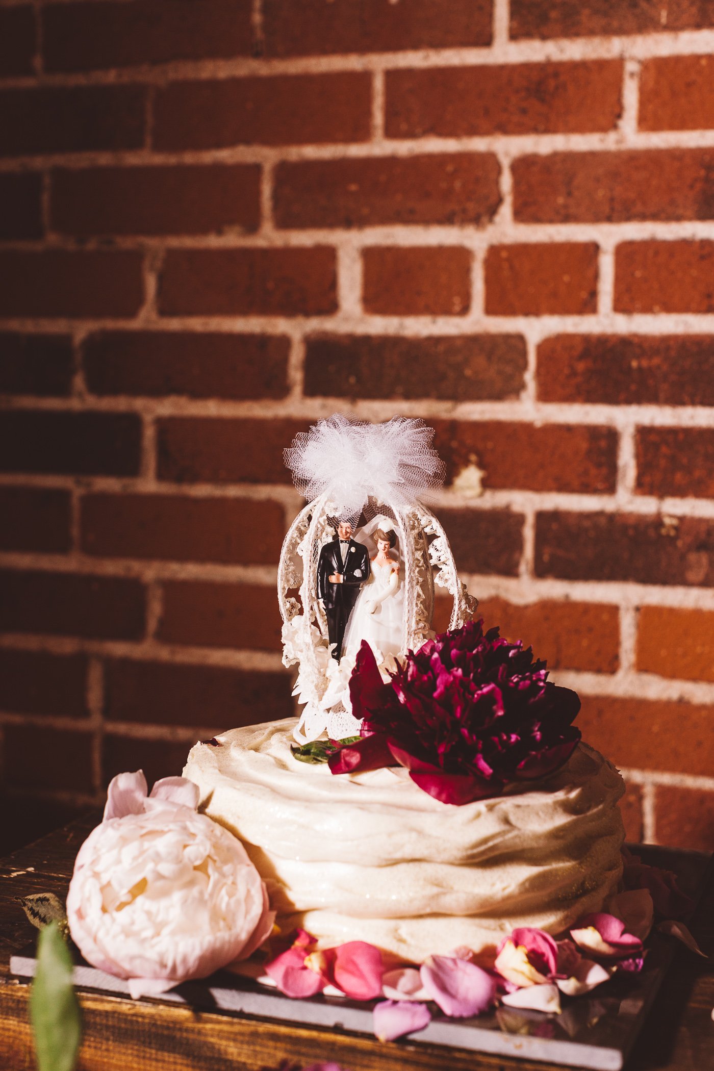 Wedding cake decorated with pink rose petals and a bride and groom topper inside a lantern, with a brick wall in the background.
