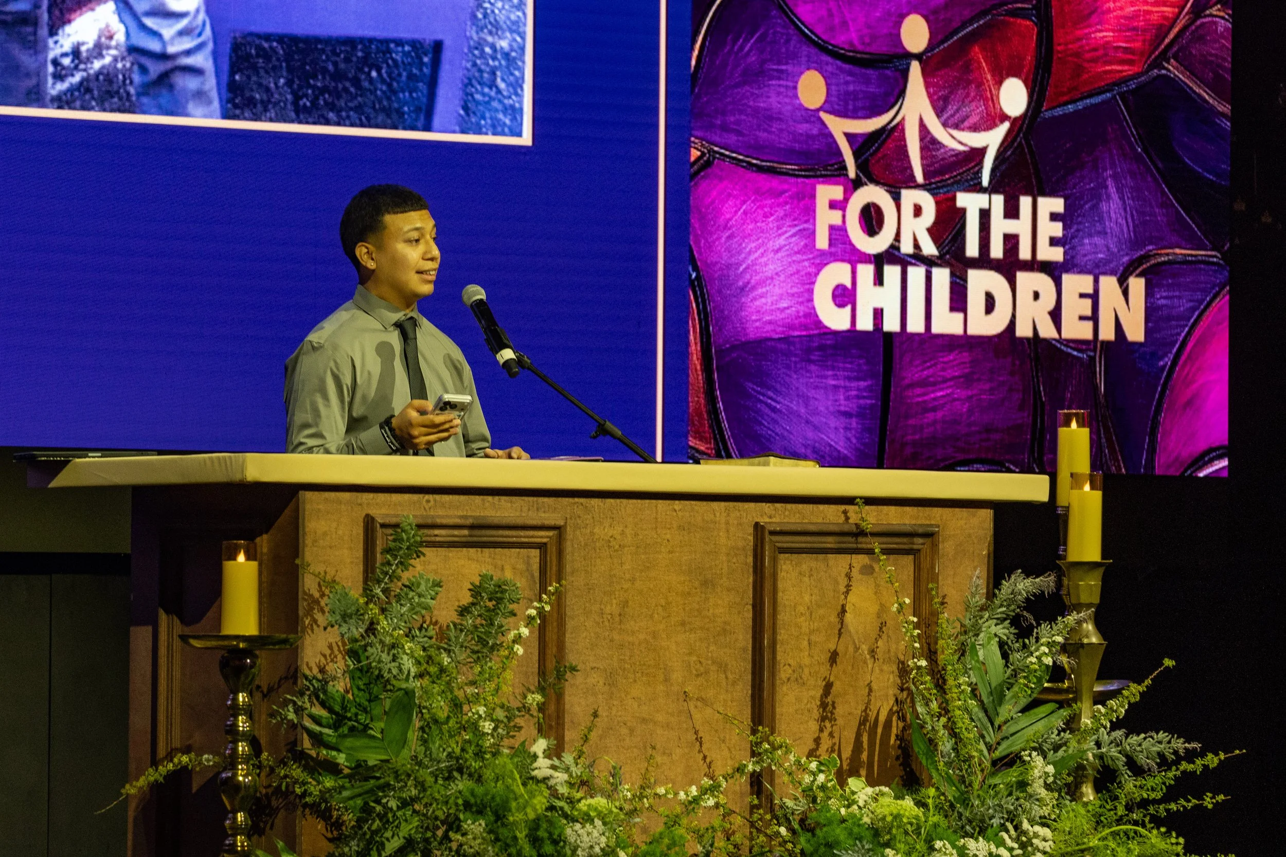 A person speaking at a podium during a event with a colorful backdrop that has the words 'For the Children' and abstract artwork, with greenery and candles in the foreground.