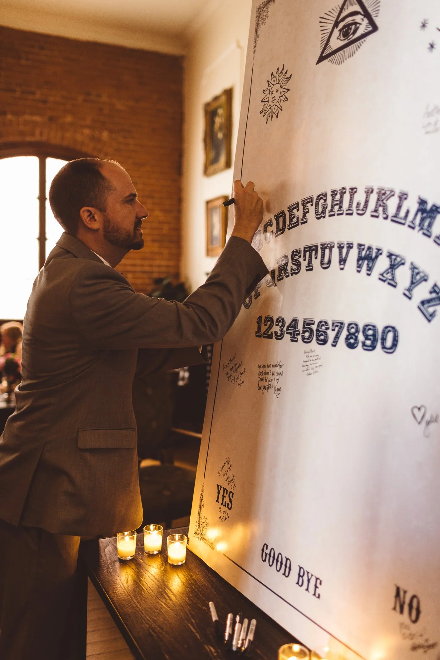 Man in a suit writing on a large white poster with black ink near candles on a dark wooden table, and framed pictures on a brick wall.