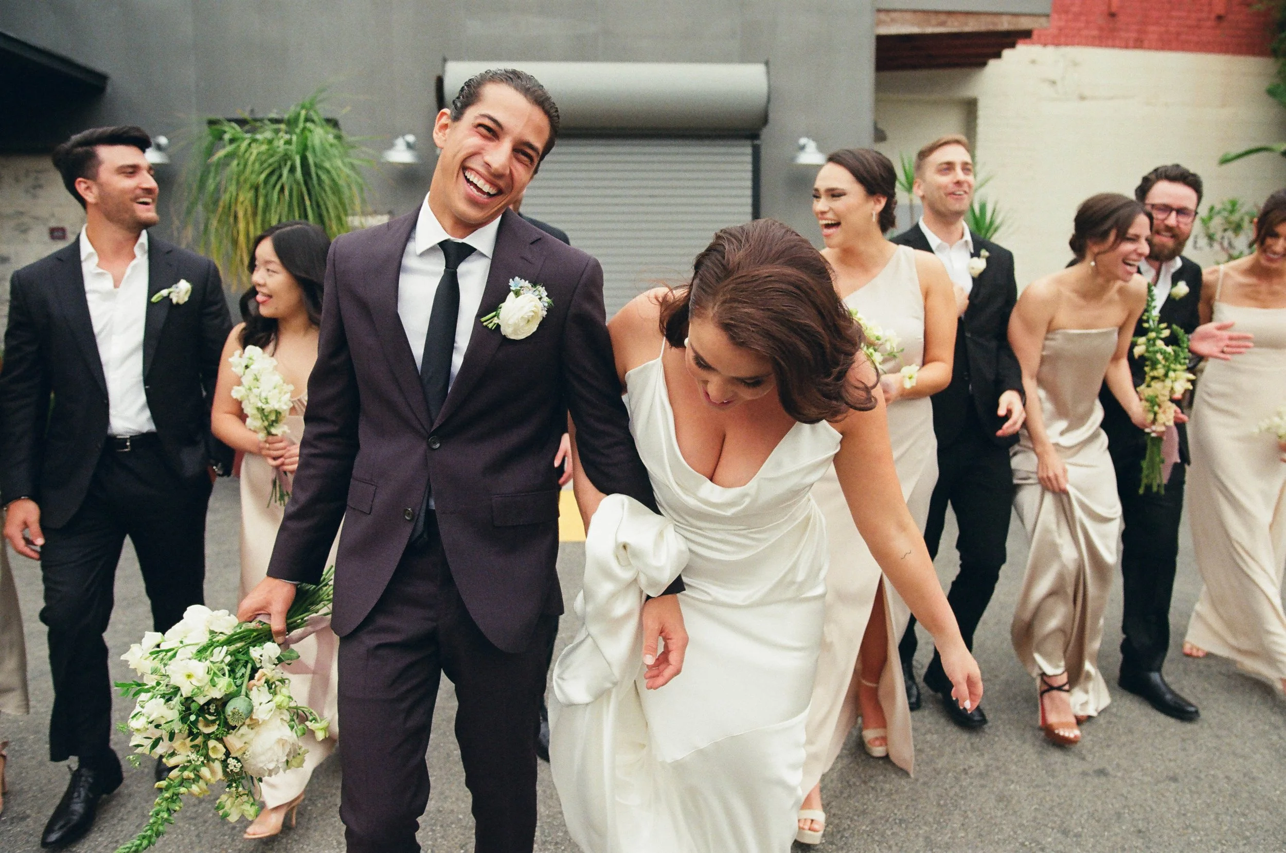 A group of smiling people dressed in formal wedding attire, walking outside in an urban area, with a man in a dark suit and a woman in a white wedding dress in the forefront.