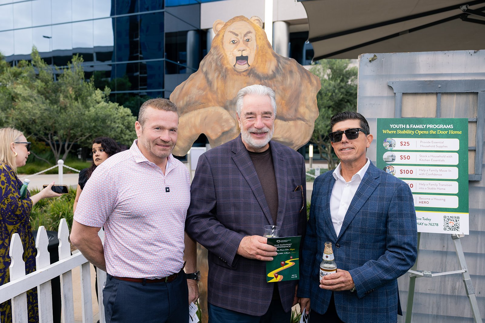 Three men standing outdoors, smiling at the camera at a community event; behind them, a large lion figure with a human face and a sign listing youth and family program donations.