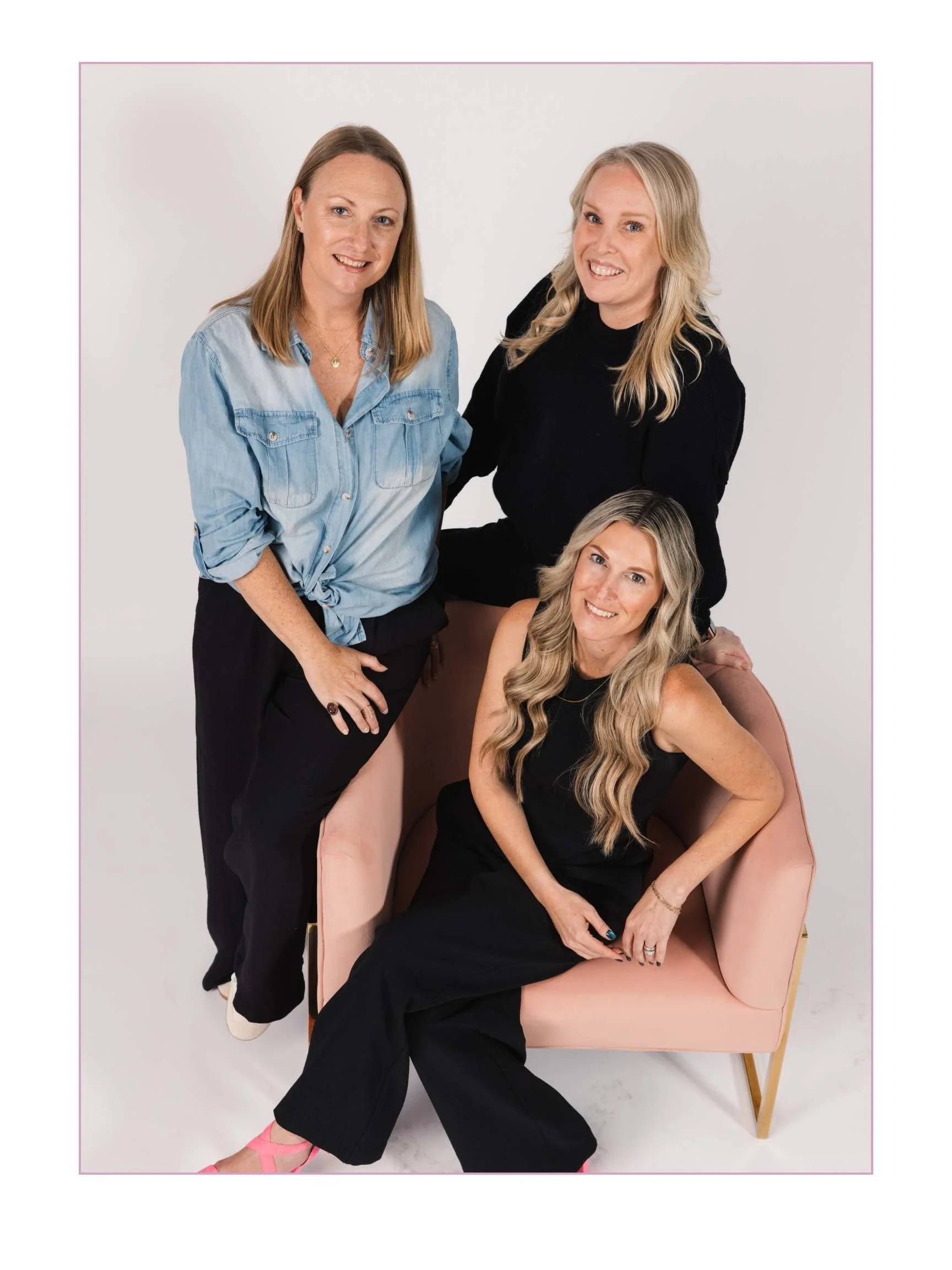 Four women posing together, one sitting in a pink armchair and three standing, against a white background.