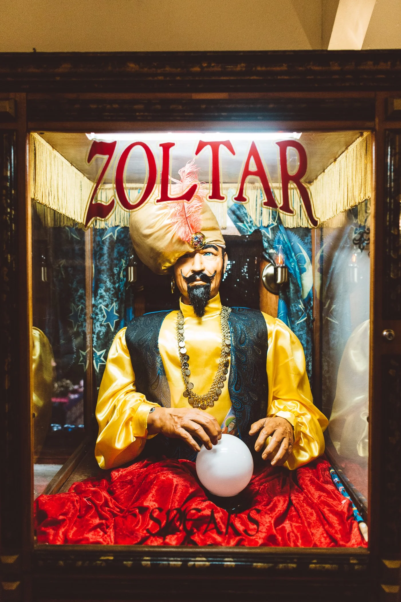A figure dressed as a fortune teller with a turban, gold robes, and jewelry, sitting inside a glass booth labeled 'ZOLTAR'. The figure holds a crystal ball in their hands and sits on a red velvet seat.