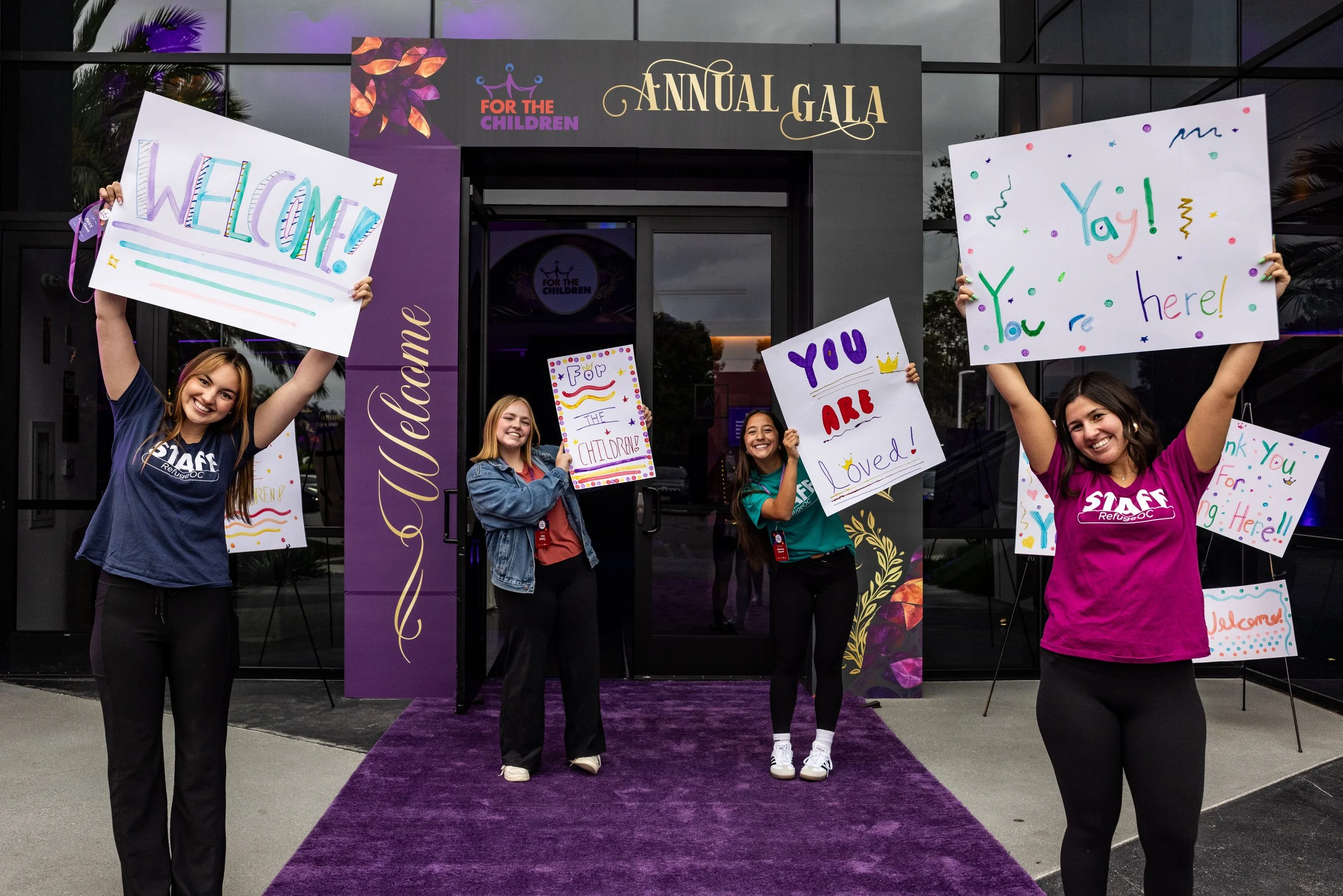 Four young women standing on a purple carpet outside a building, holding colorful signs with welcoming messages for a children's event, with a large sign above for an annual gala for the organization 'Refuge OC' and 'For the Children'.