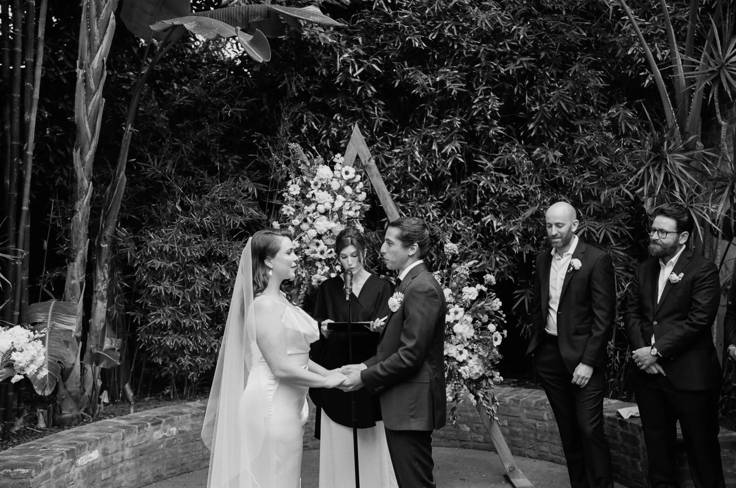 Black and white photo of a wedding ceremony with a couple holding hands and exchanging vows, surrounded by an officiant and wedding party, with lush greenery and flowers in the background.