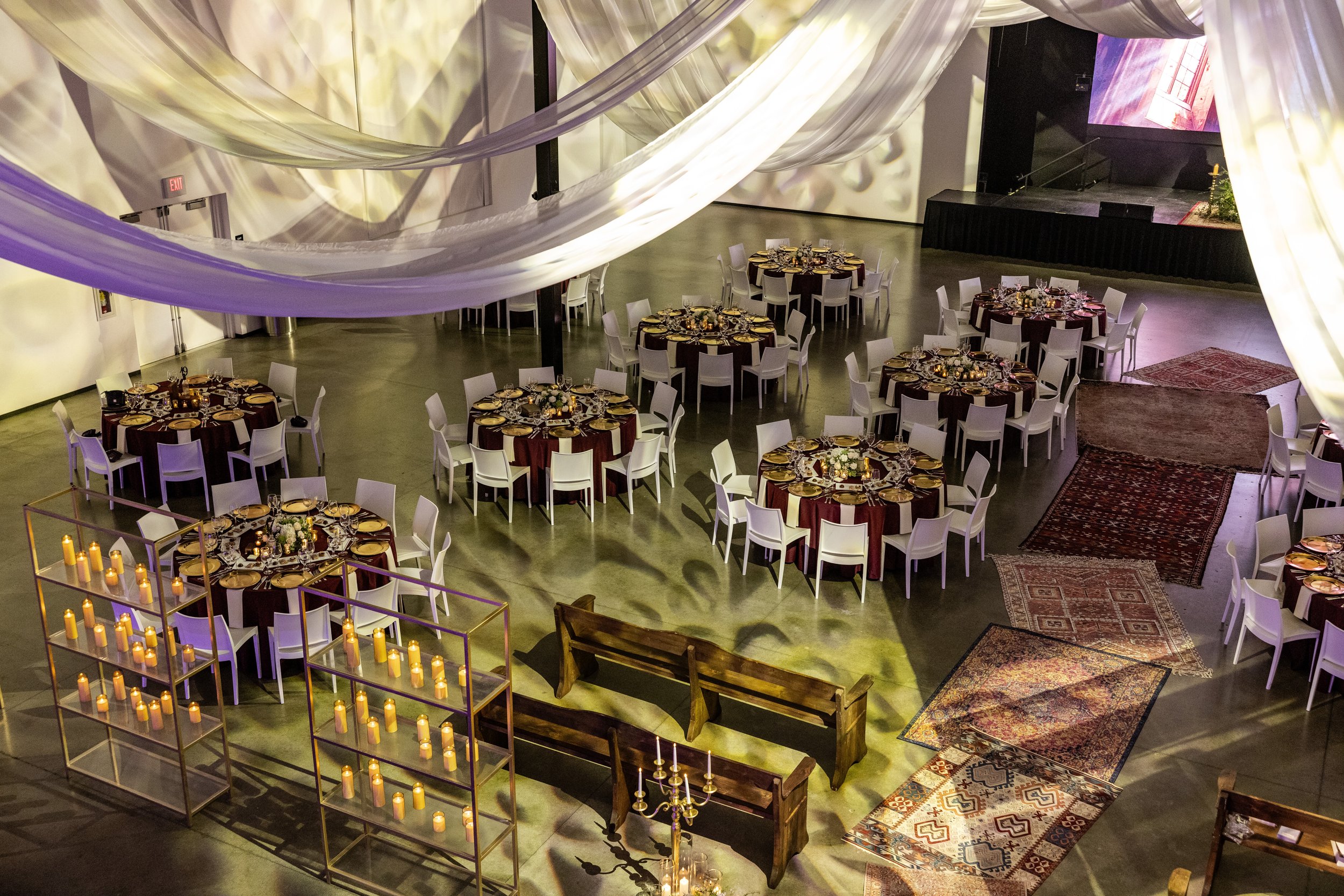 Event space decorated with draped fabric, round tables with burgundy tablecloths and white chairs, candles, and floral centerpieces. There are rugs on the floor and a small stage with a large screen. Candles are arranged in a gold-framed shelf near a