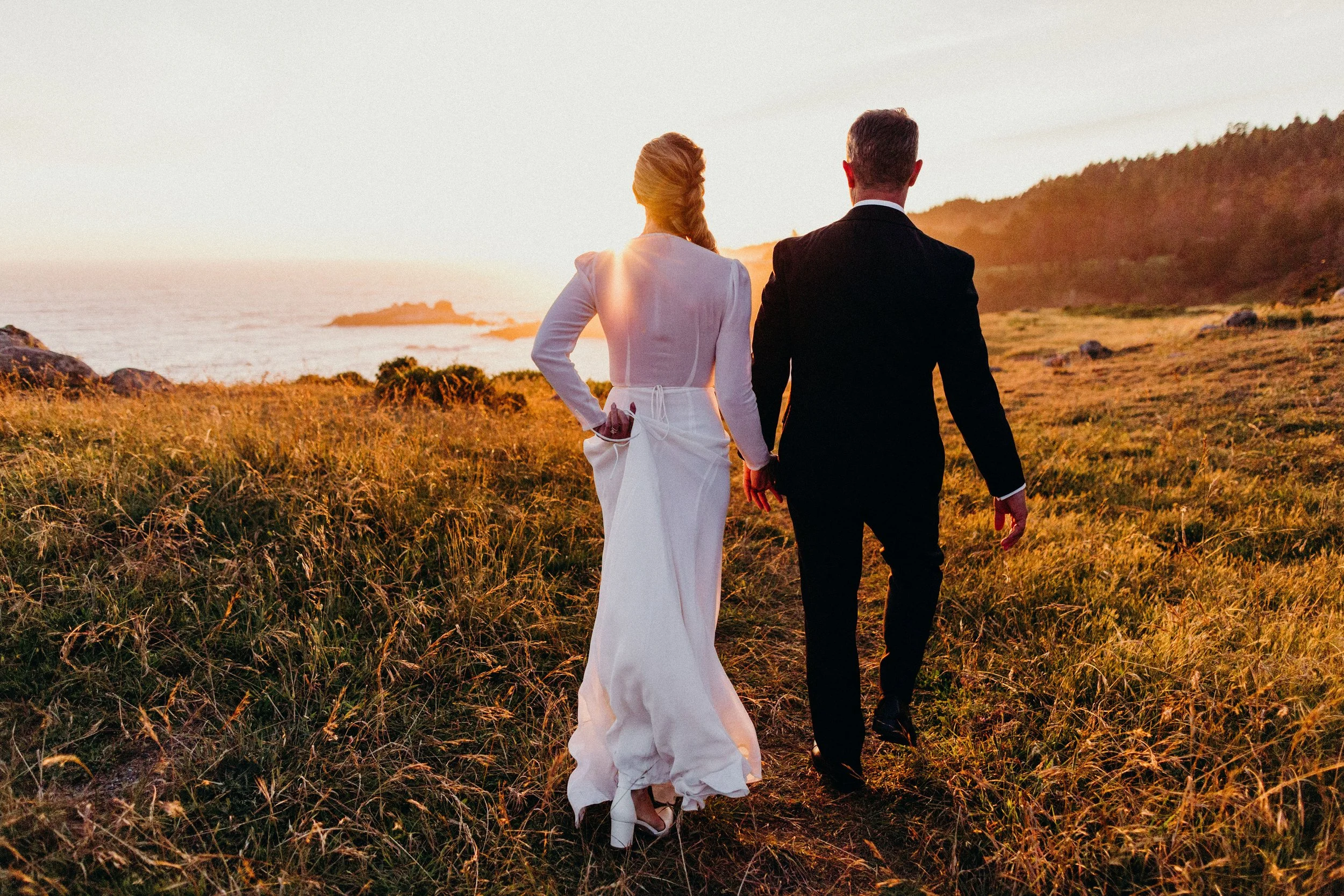 A couple walking hand in hand along a grassy path at sunset near the coast. Amy + Dave, Timber Cove 