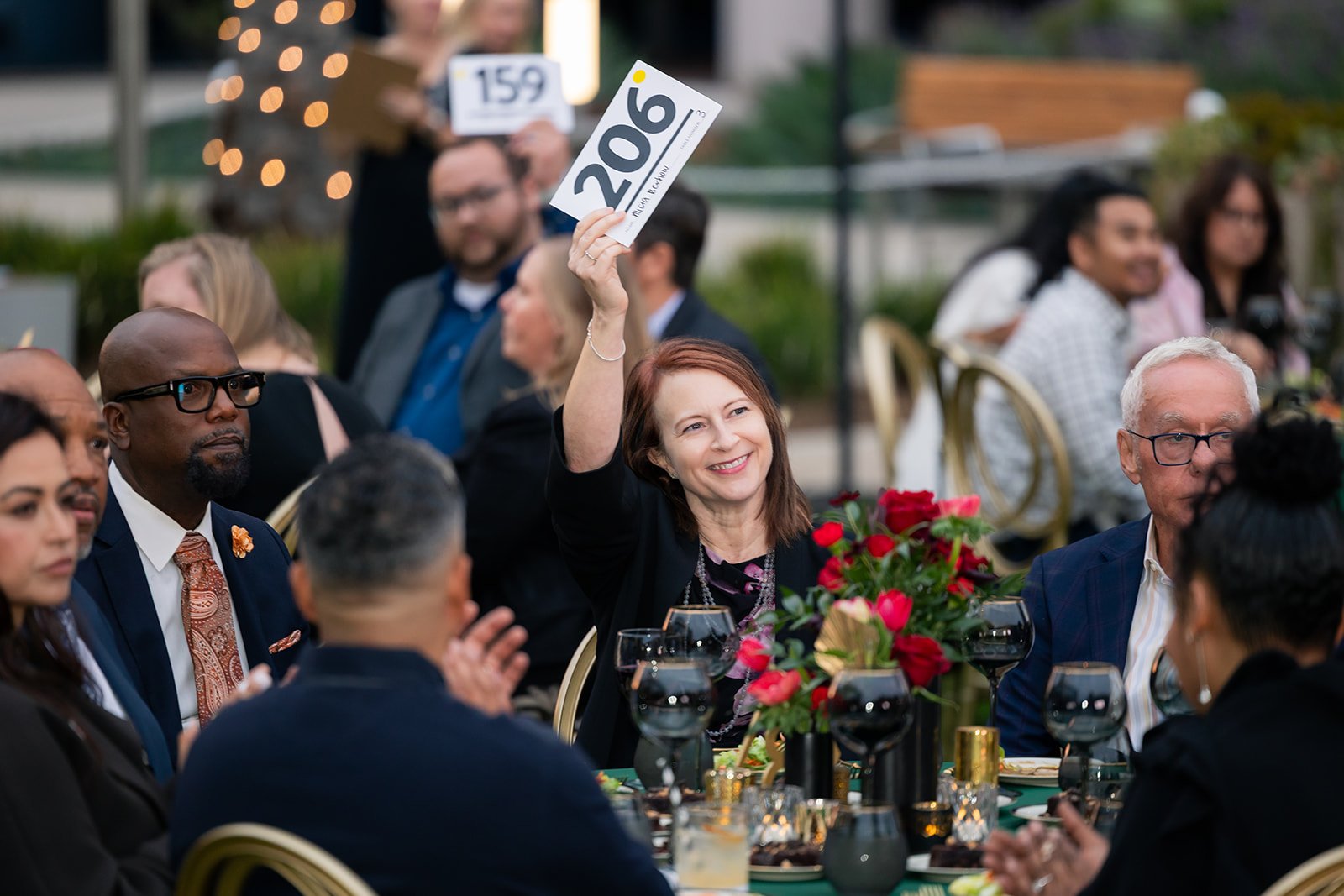 A group of people seated at a dinner table outdoors, with one woman smiling and holding up a card that reads '206'. The table is decorated with a bouquet of pink and red flowers and glasses of red wine.