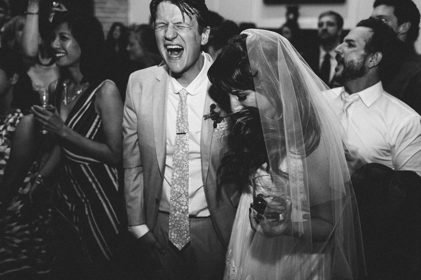 Black and white photo of a wedding celebration with a group of people. A man in a light suit and tie is singing or shouting with joy. A woman in a wedding dress and veil is leaning into him, holding a glass, smiling with her eyes closed. The crowd ar