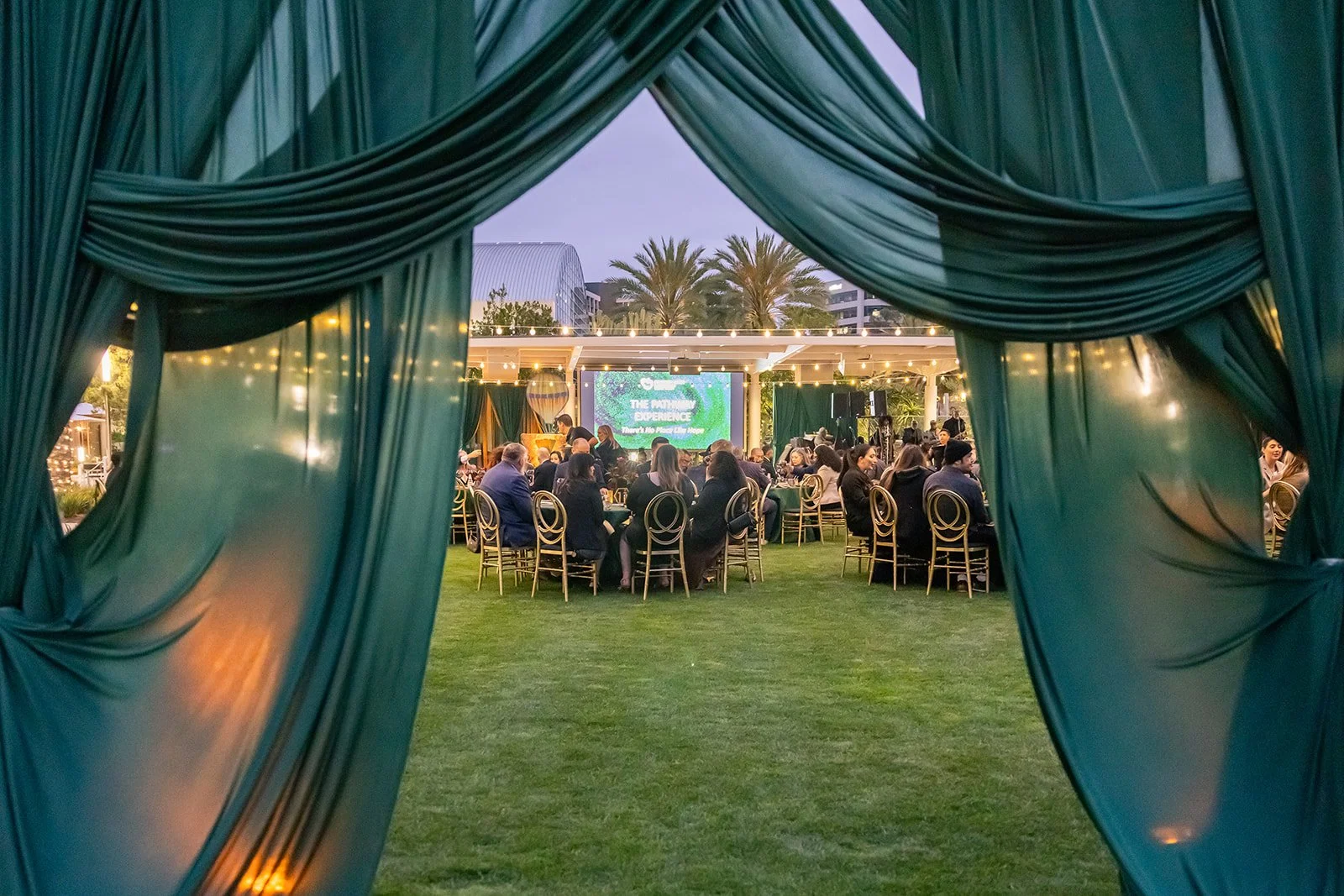 Outdoor event with round tables and chairs, green curtains framing the scene, and a large screen displaying a presentation, in a garden setting with palm trees in the background.