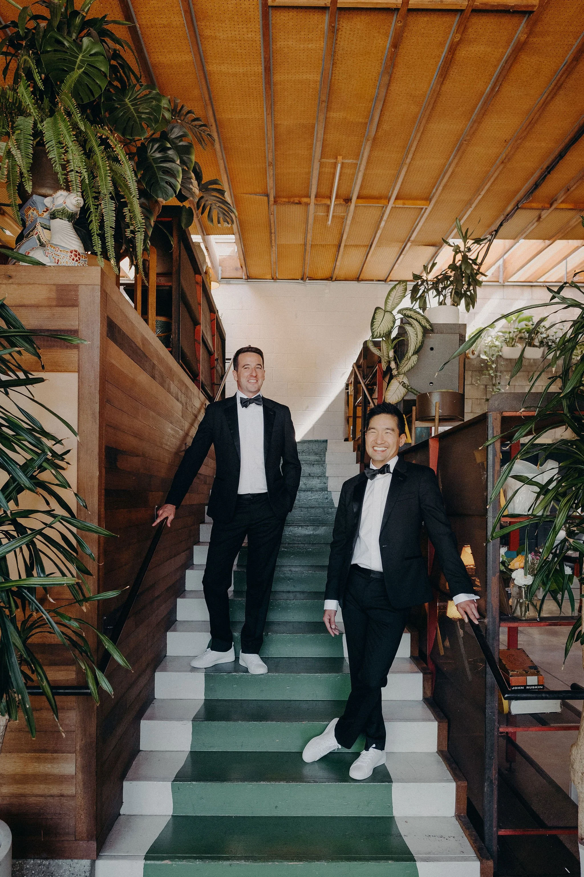 Two men in tuxedos standing on a green and white staircase, surrounded by indoor plants and wood-paneled walls, smiling at the camera.