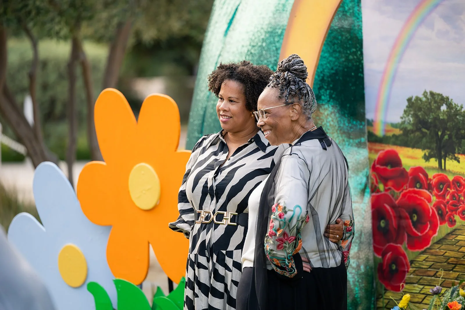Two women stand close together in front of colorful flower and garden-themed decorations, smiling and sharing a moment outdoors.