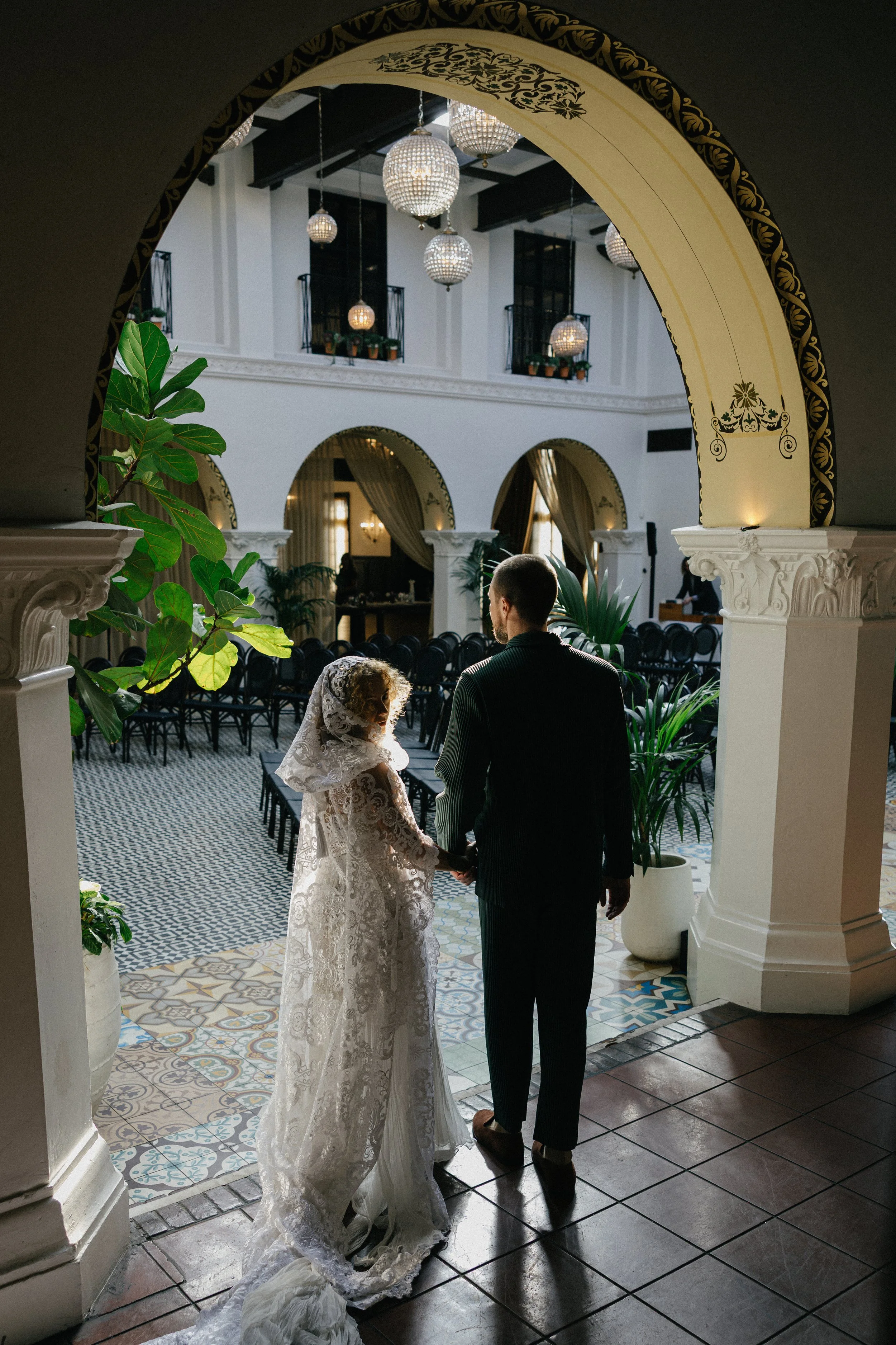 A bride and groom hold hands and stand together inside a decorated venue. The bride is wearing a lace wedding gown and veil, while the groom is dressed in a dark suit. They are seen from behind as they prepare to get married or have just been married, with ornate chandeliers hanging from the ceiling and potted plants surrounding them.