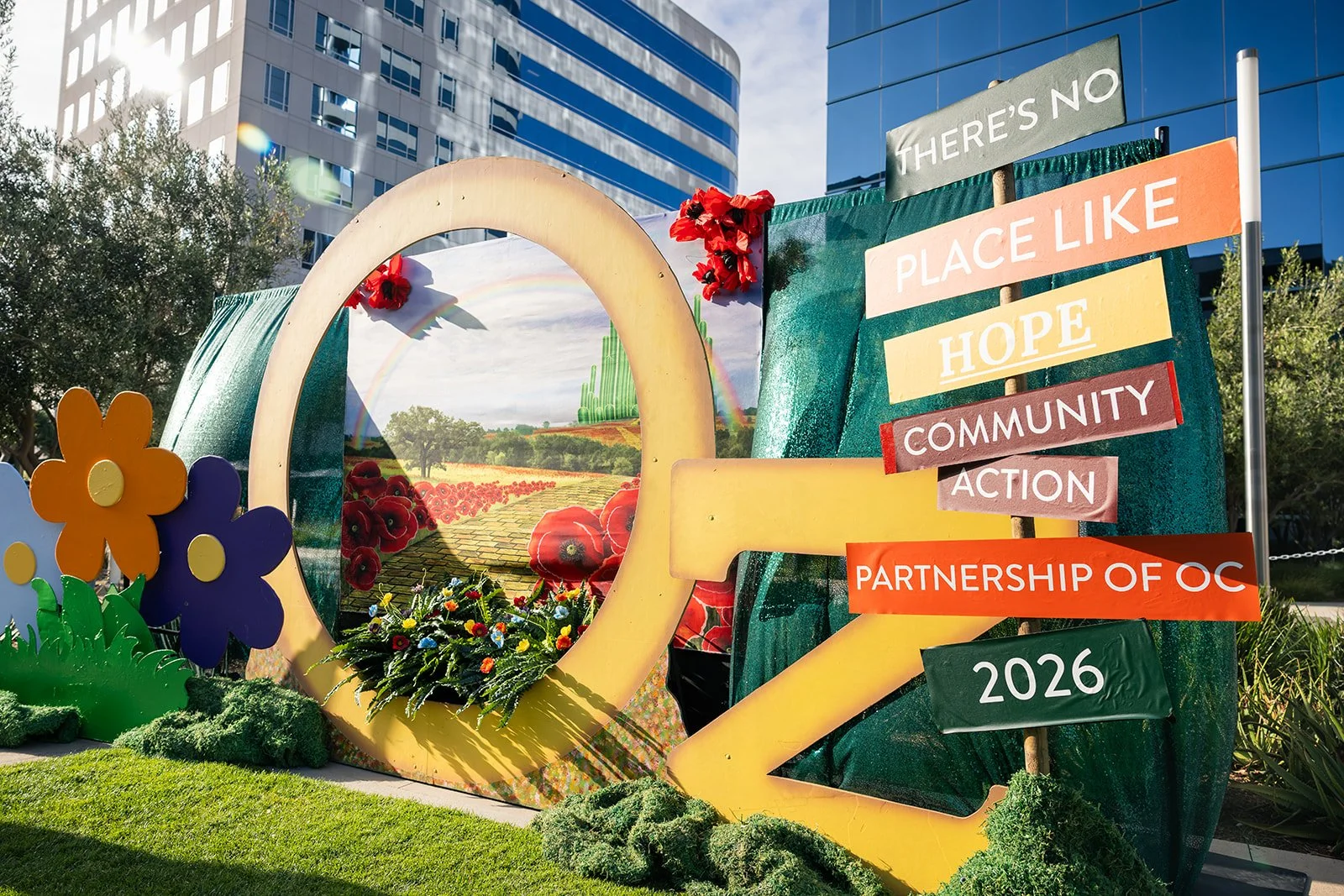 Outdoor art installation with colorful flowers, a circular frame, and a painted landscape featuring a rainbow, poppies, and greenery. A sign with words like 'Place Like Hope' and 'Community Action' and the year 2026 is displayed next to the scene.