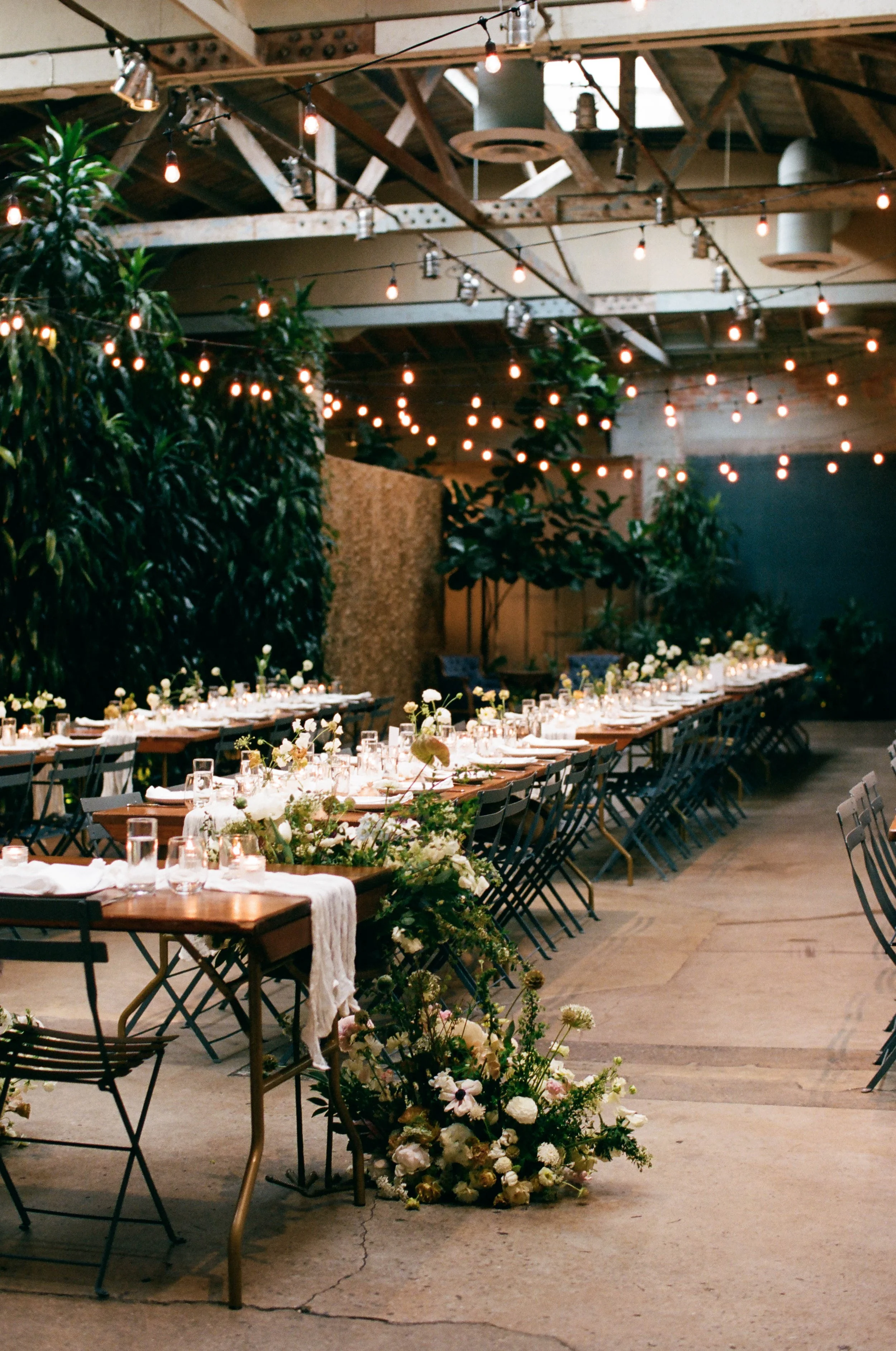 Long banquet table decorated with flowers and candles, set in an industrial-style venue with string lights hanging from wooden beams, and lush plants and flowers along the sides.