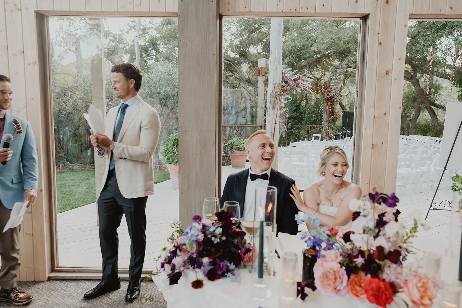 Wedding reception with two seated guests laughing, one man in a tuxedo and one woman in a white dress, with floral arrangements on the table. Two men are standing outside a large glass window, one holding a microphone and the other reading a paper.