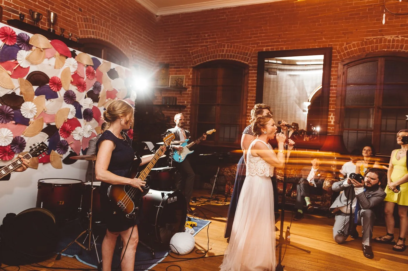 A wedding reception with a live band performing indoors with brick walls and large windows. The bride is singing into a microphone, and the band members are playing guitars and drums. Guests are watching, with some taking photos.