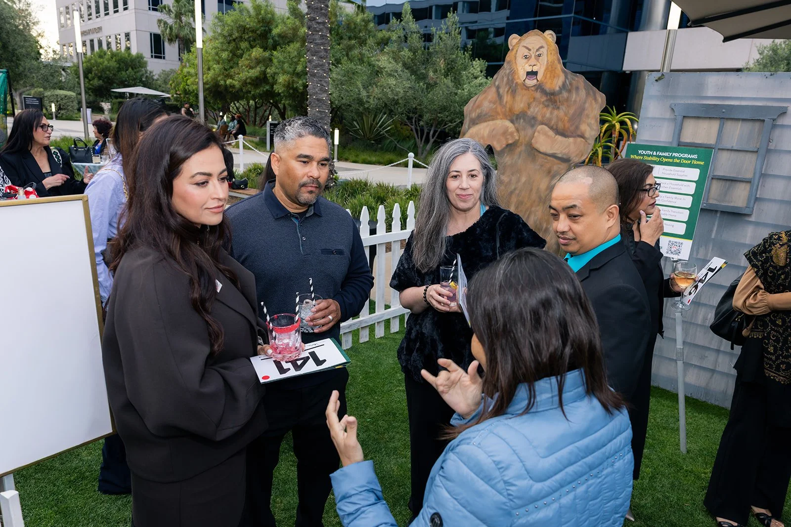 A group of six diverse people at an outdoor event, engaged in conversation, holding drinks, with a wooden fence and trees in the background, and a large lion cutout statue nearby.
