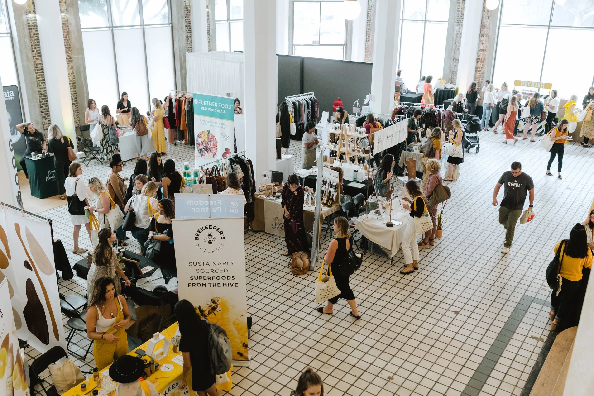 A crowded indoor market or fair with various vendor booths selling clothing, jewelry, and food. People are browsing, shopping, and walking around in a spacious, well-lit area with large windows and tiled floors.