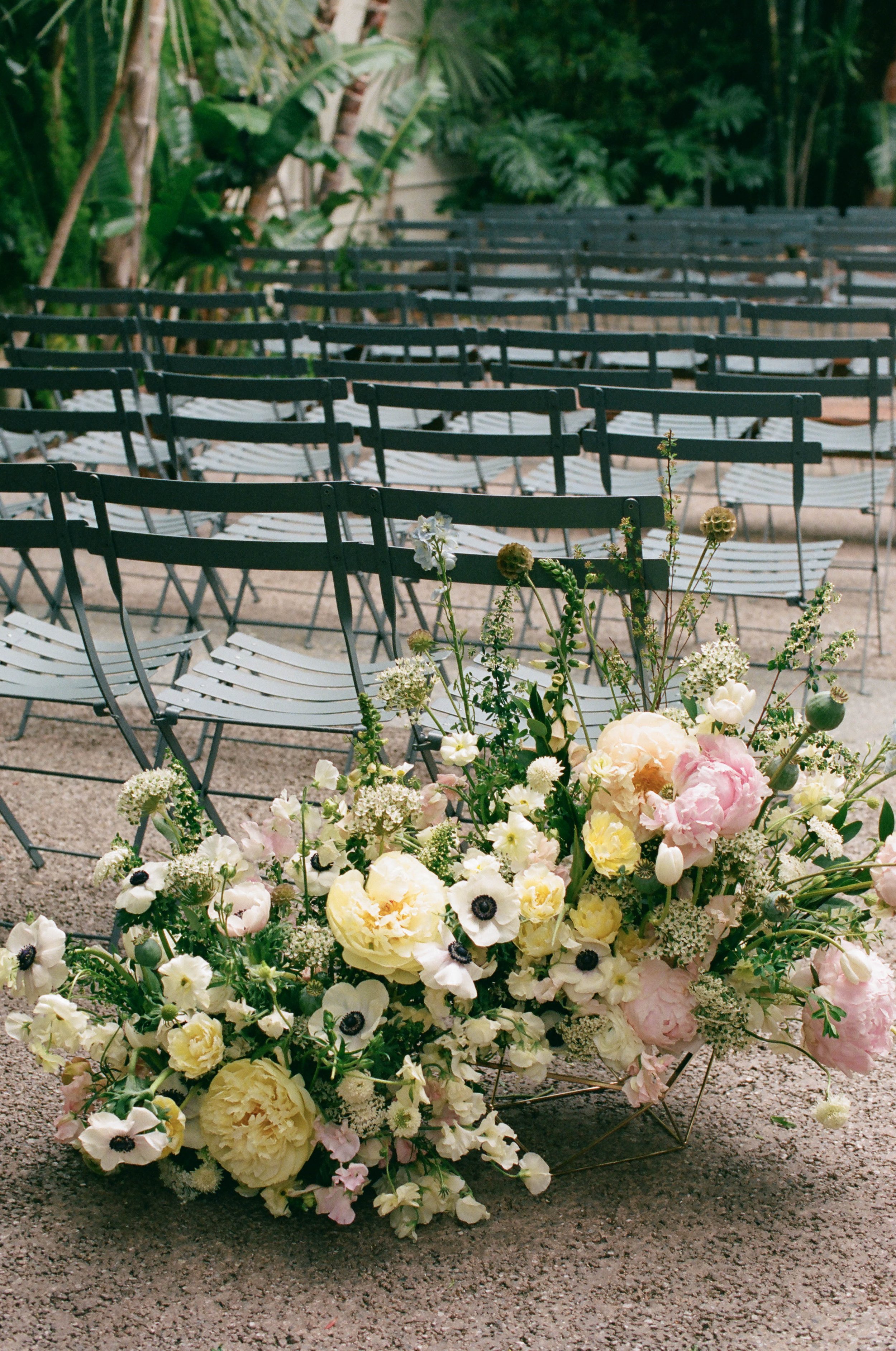 A large bouquet of pastel-colored flowers, including white, pink, and yellow blooms, arranged on the ground outdoors in front of green foliage. In the background, there are several rows of empty black metal chairs.