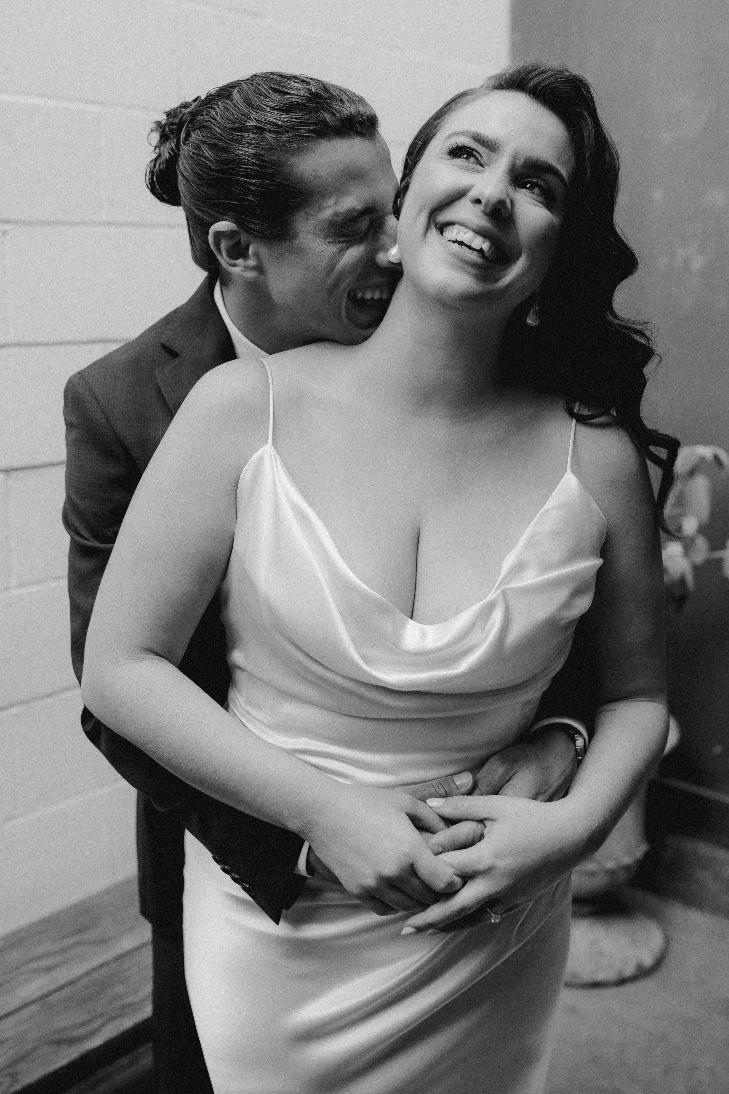 A smiling woman in a satin dress is embraced by a man in a suit, who is nuzzling her neck and whispering in her ear.