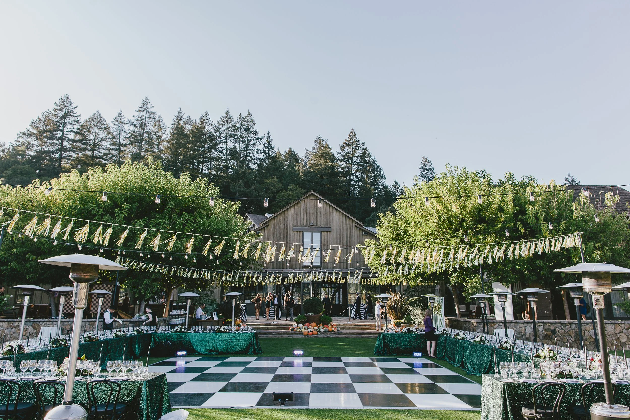 A decorated outdoor event space with a black and white checkered dance floor, surrounded by greenery, string lights, and tables with umbrellas, set against a backdrop of trees and a small building.