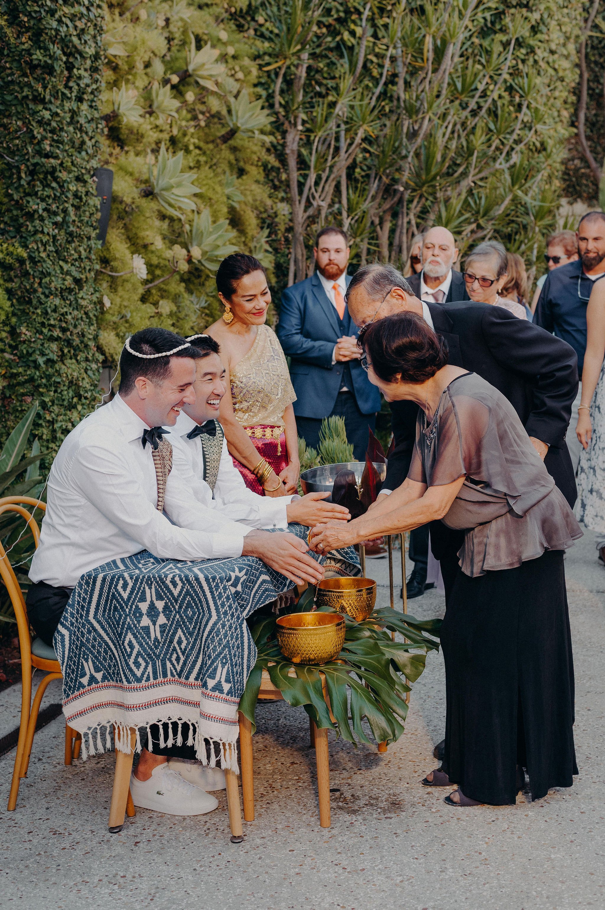 People participating in a wedding ceremony outdoors, with the bride and groom seated, and guests standing around, exchanging respectful bows. The scene is surrounded by lush greenery and decorated with gold bowls and tropical leaves.
Cliff + Warren