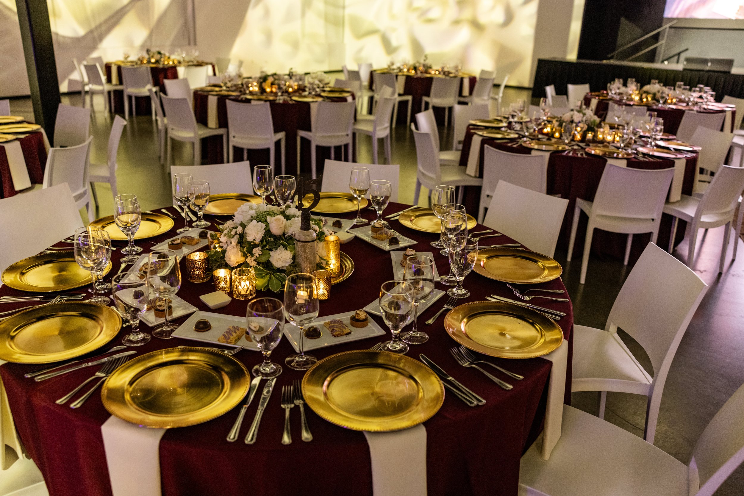 A banquet hall with round tables decorated with white and burgundy tablecloths, gold chargers, and elegant glassware. The tables feature floral centerpieces with white flowers and candles, creating a festive atmosphere.